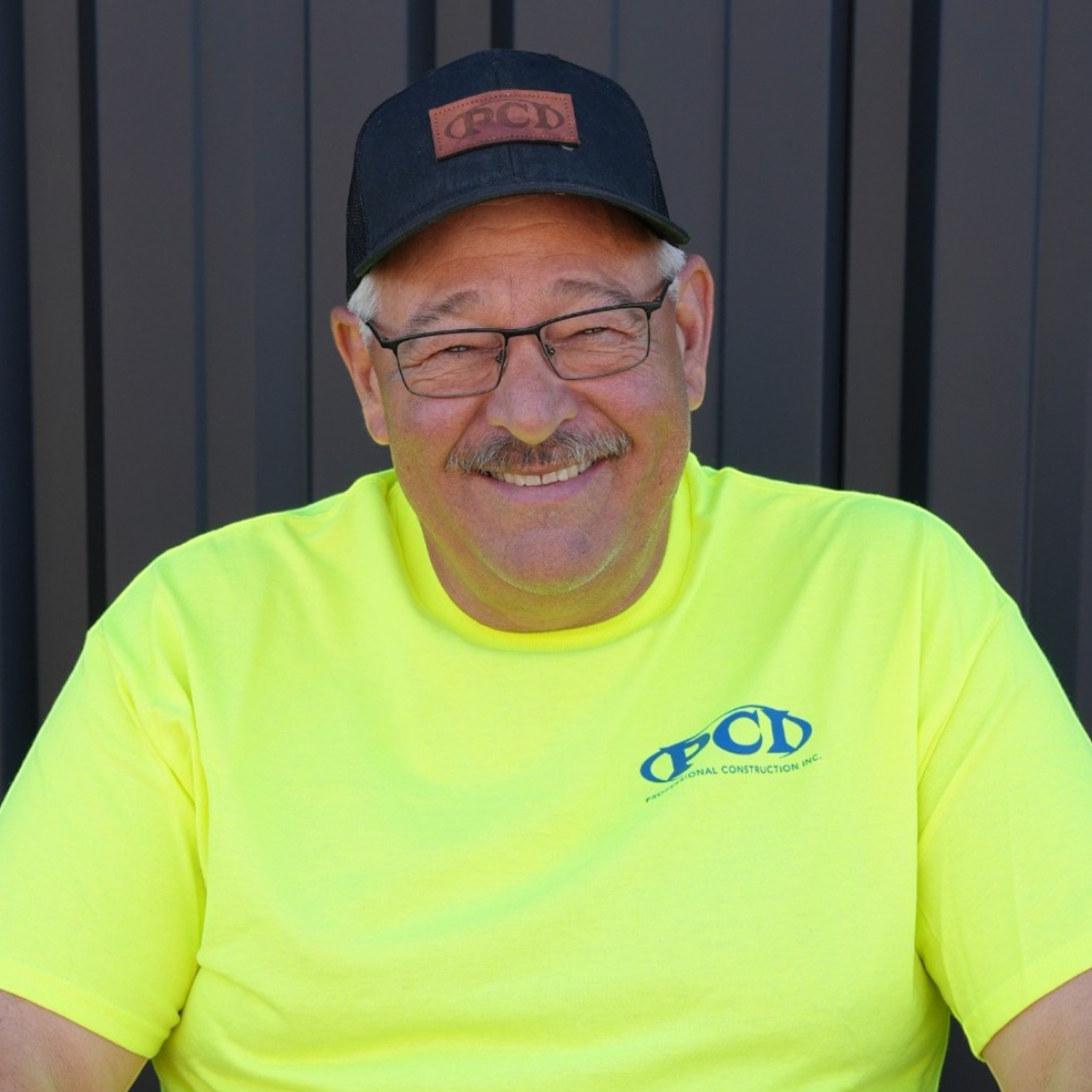 A middle-aged man with glasses, a mustache, and a goatee wearing a gray Columbia cap and a black polo shirt with a logo for PCD Professional Construction Inc. standing in front of a gray metal wall.