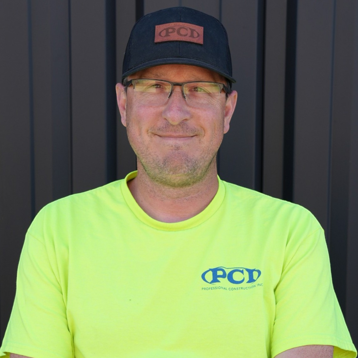 A man wearing glasses, a black cap with white logo, and a black polo shirt with a logo that says 'PCD Professional Construction Inc.' standing in front of a dark gray, metal-paneled background.