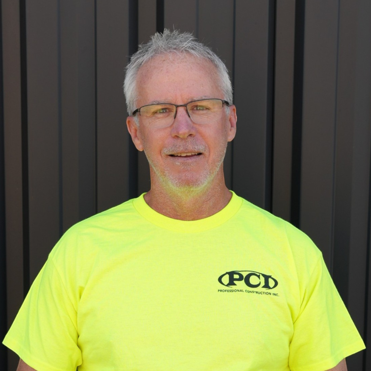 Man wearing glasses, a cap, and a black polo shirt with the logo of Professional Construction Inc. standing in front of a dark gray wall.