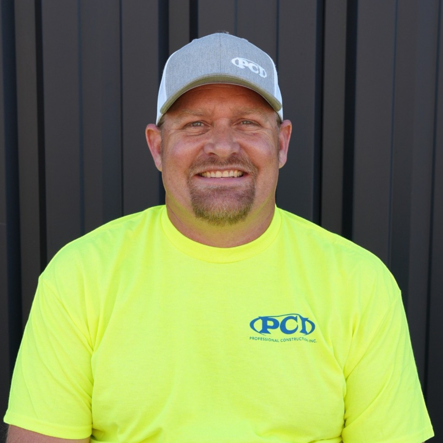 A smiling man wearing a black Polo shirt and a black and white cap, standing in front of a dark grey metal wall.