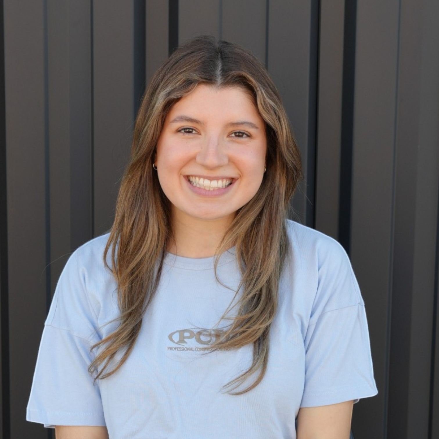 A smiling young woman with shoulder-length light brown hair wearing a black t-shirt that reads 'Professional Construction Inc. Est. 2004' standing in front of a dark exterior wall.
