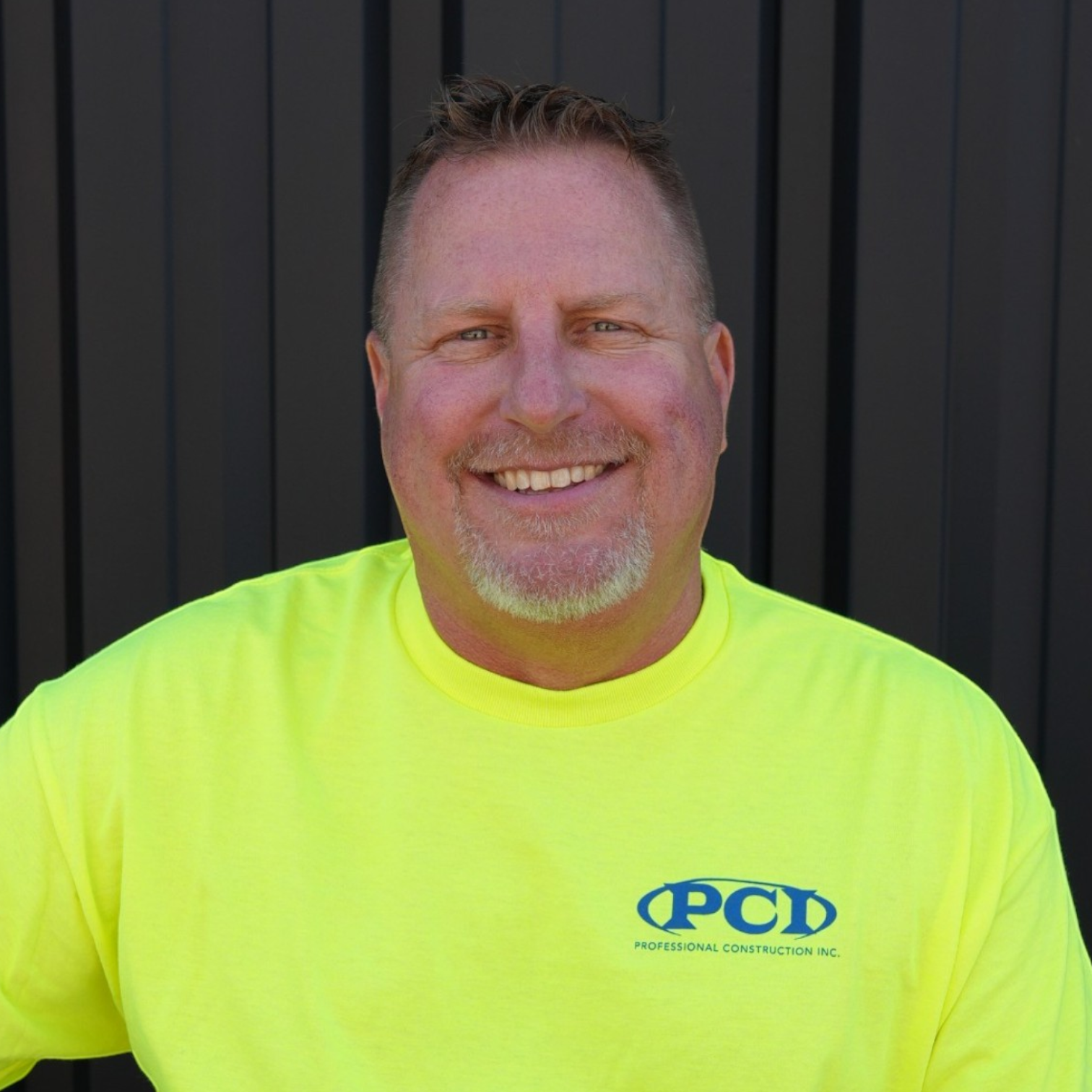 A man wearing a black baseball cap and a black t-shirt with a logo that reads 'PCD' and 'Professional Construction Inc.', standing in front of a black metal fence.
