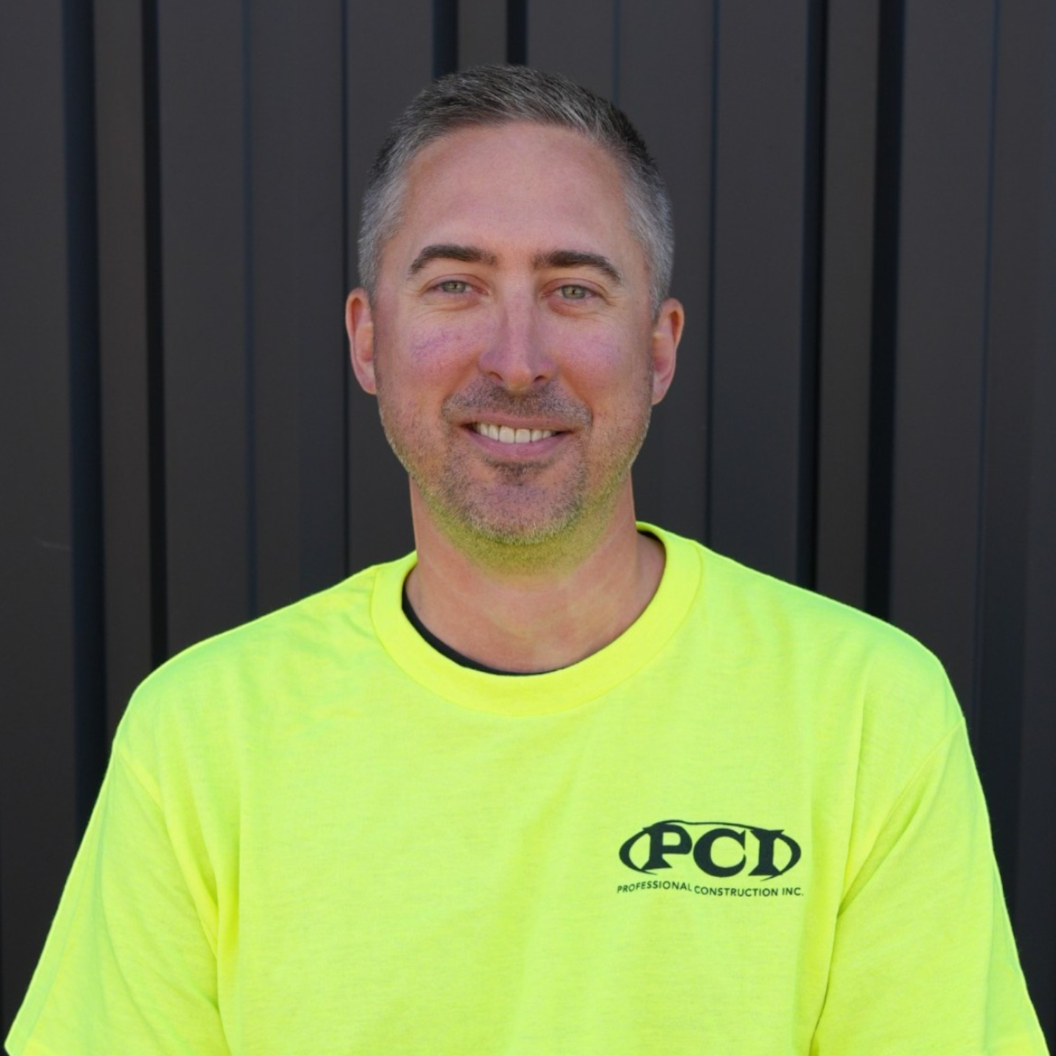 A smiling man with short gray hair and blue eyes standing in front of a wooden fence, wearing a gray t-shirt that reads "Professional Construction Inc. Est. 2004".