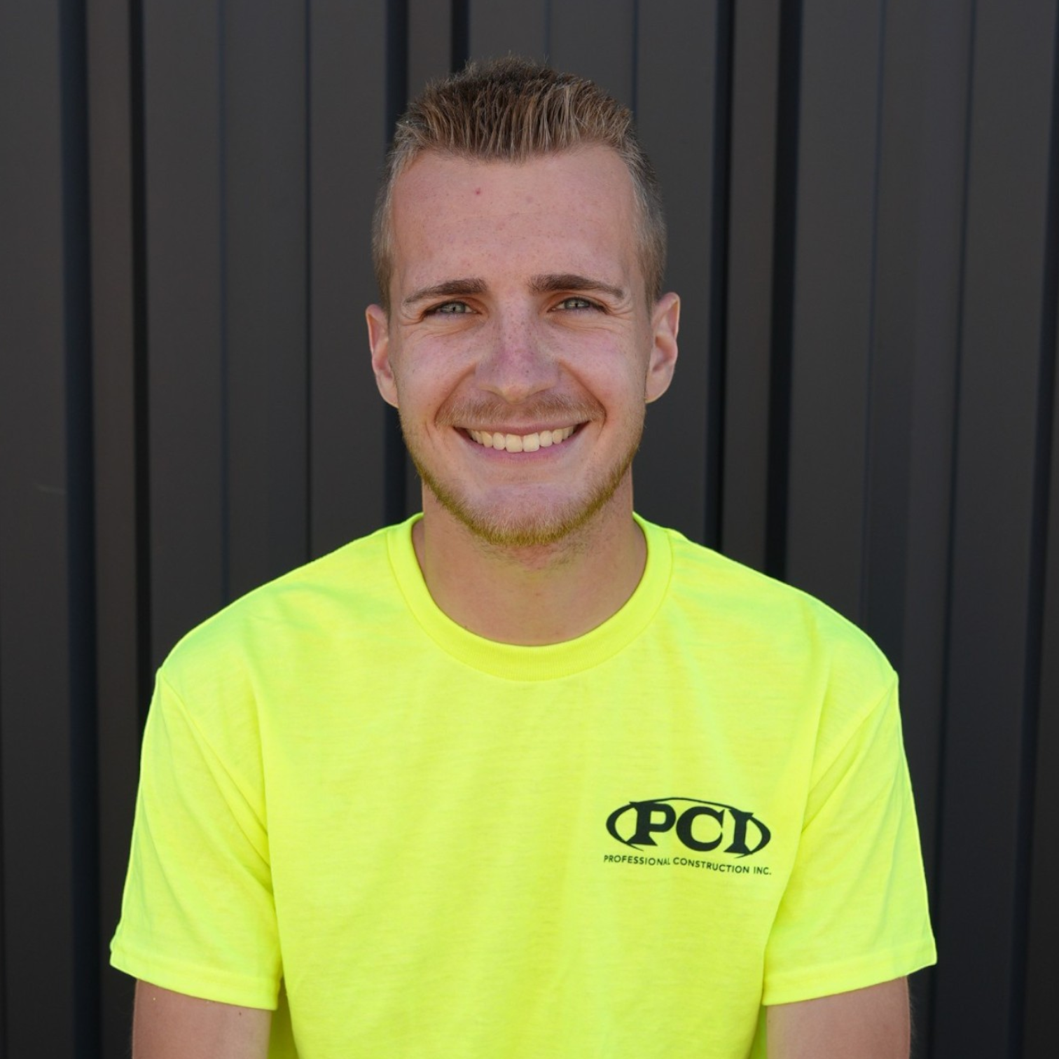 A young man smiling outdoors against a dark blue wall, wearing a black t-shirt with a logo that reads 'PCD Professional Construction Inc.'.