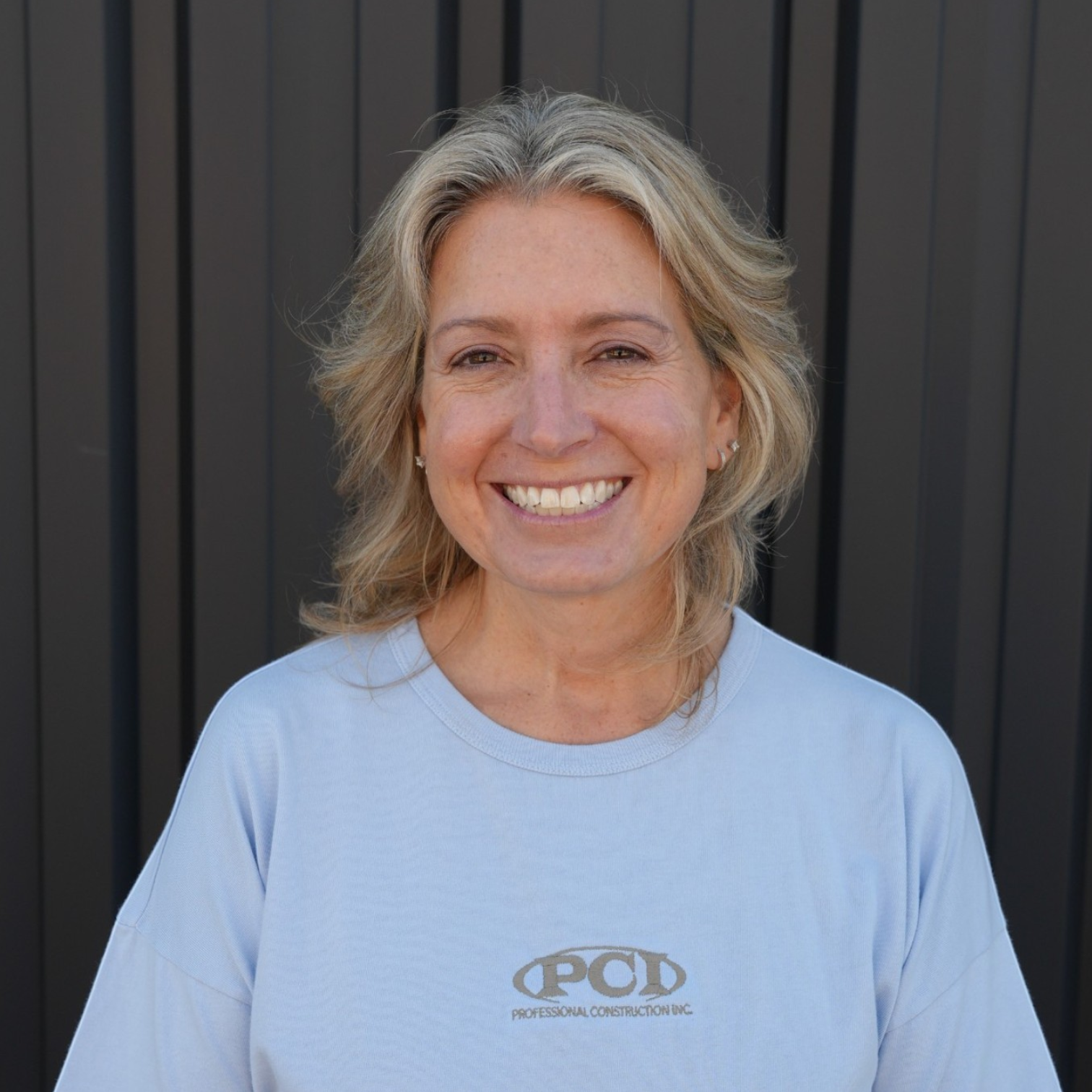 Portrait of a woman with blonde hair smiling in front of a dark wooden background.