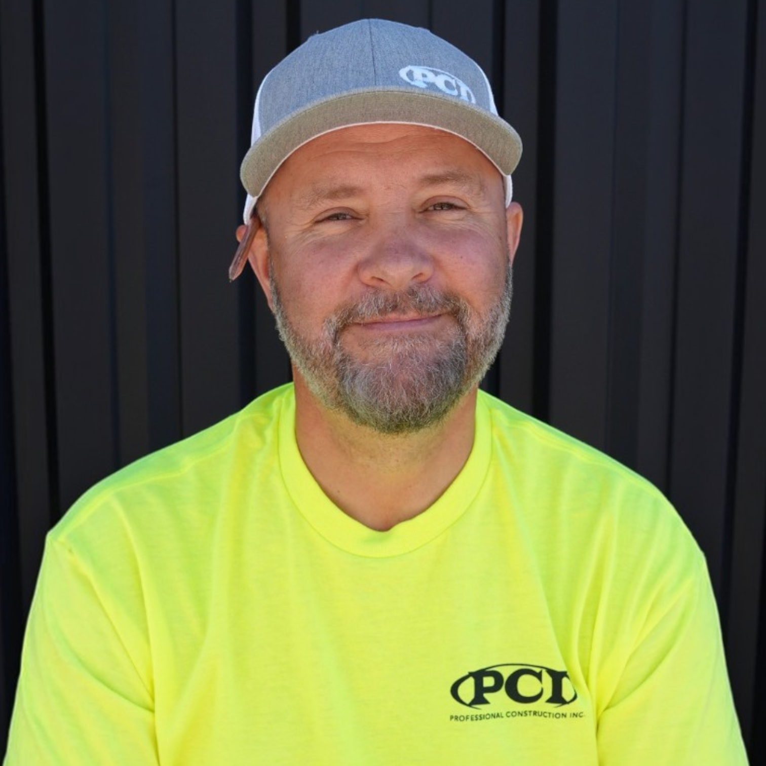 A man with a beard and short hair wearing a black PCD cap and a black T-shirt with the PCD logo, standing in front of a dark gray metal wall.