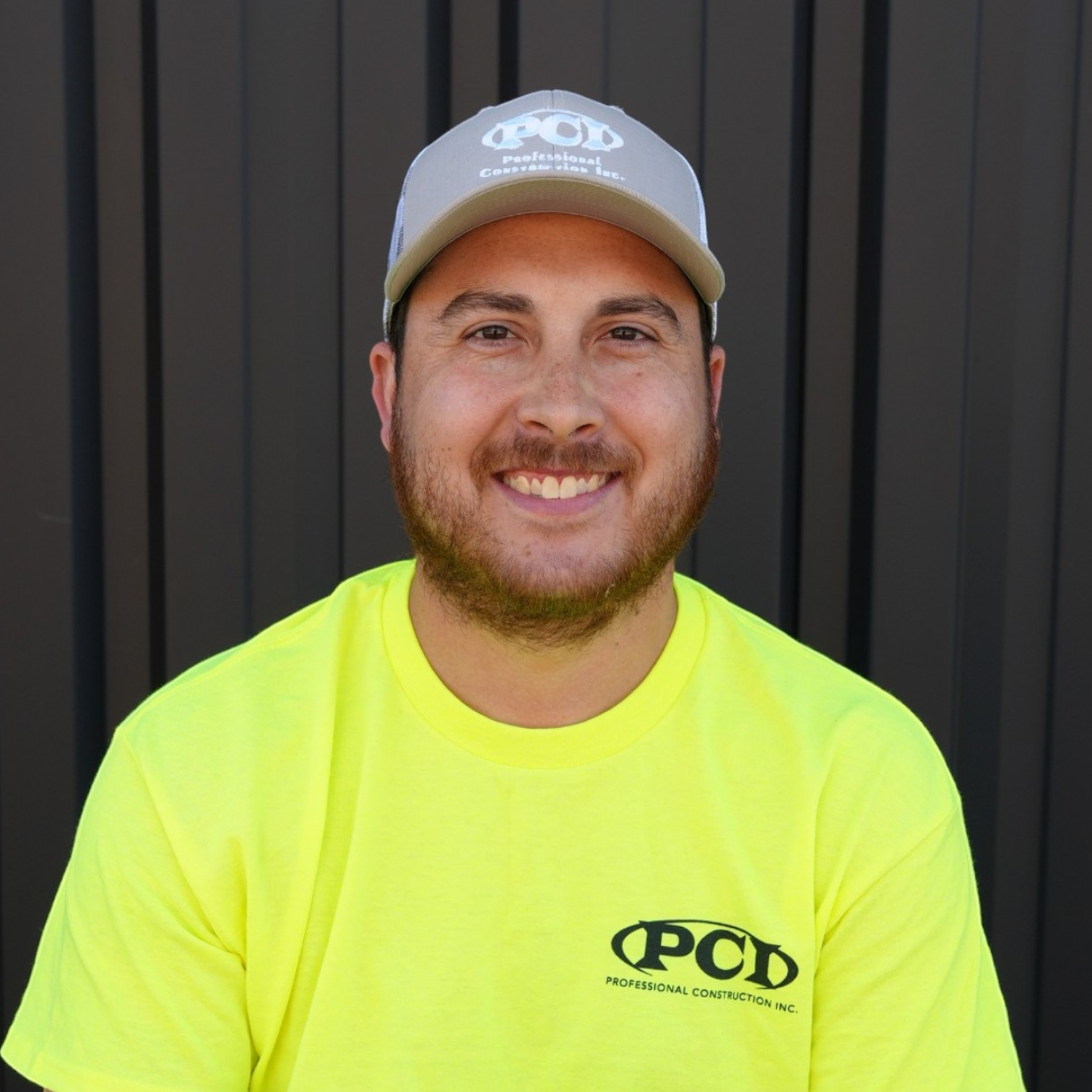 Young man with a beard smiling, wearing a camouflage baseball cap and a black T-shirt with a white logo that reads 'PCD Professional Construction Inc.' in front of a dark gray metal wall.