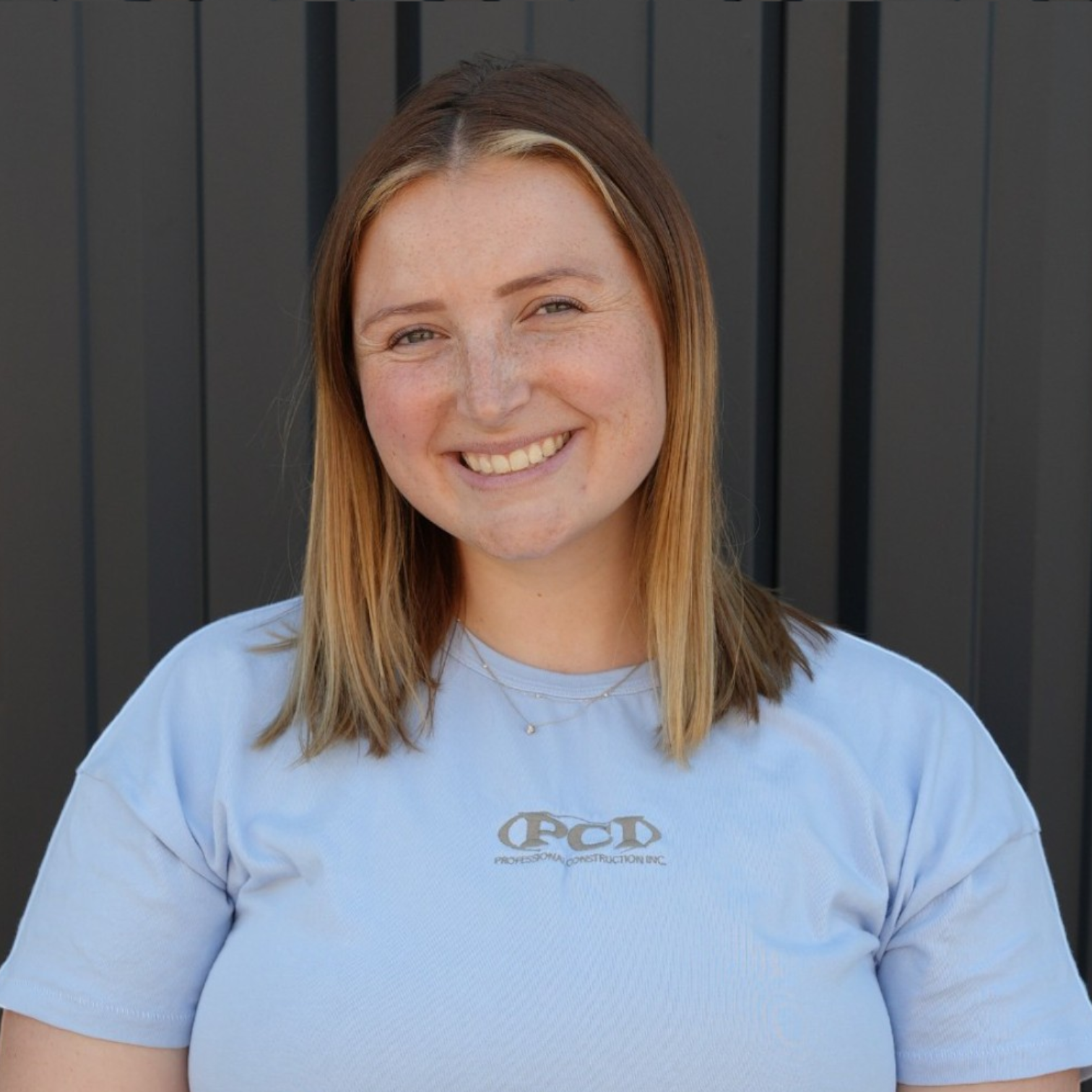 A smiling woman with blonde hair wearing a black T-shirt with a logo, standing in front of a dark blue panel background.