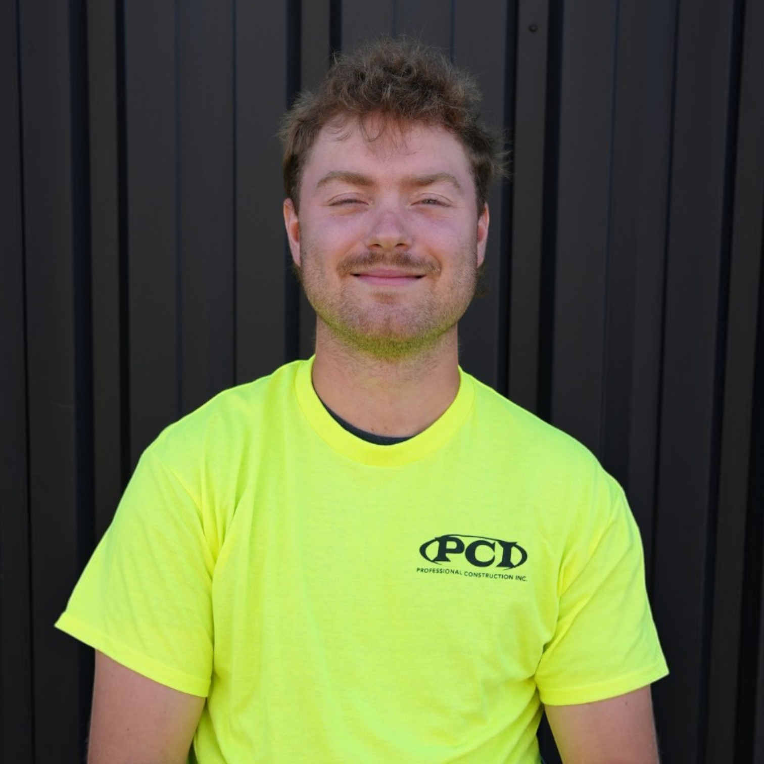 A young man with light brown hair and a mustache, smiling, wearing a black polo shirt with a logo for a construction company, standing in front of a dark gray metal wall.