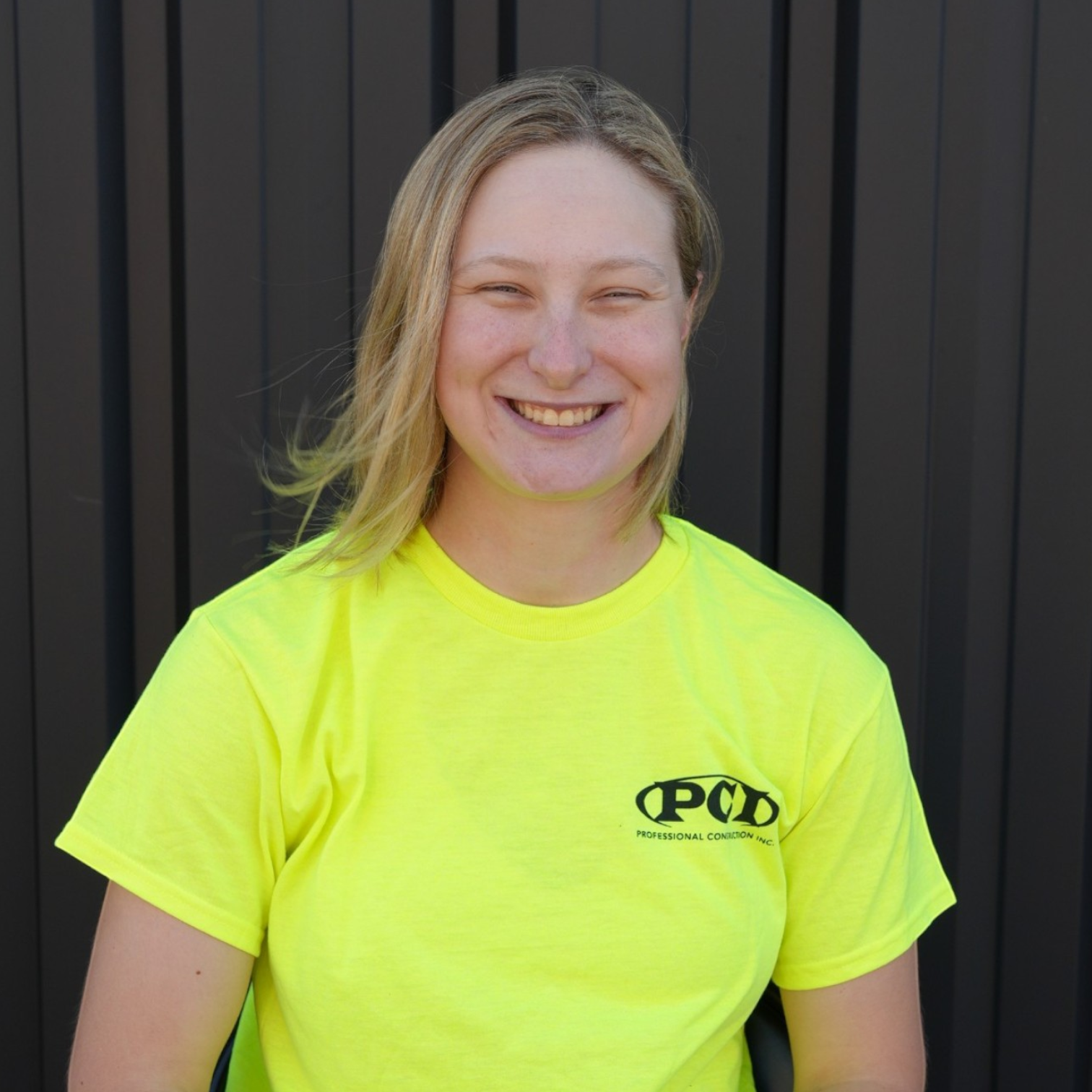 A woman with shoulder-length blonde hair smiling at the camera, wearing a black shirt with a logo that says 'PCD Professional Construction Inc.' in front of a dark gray wall.