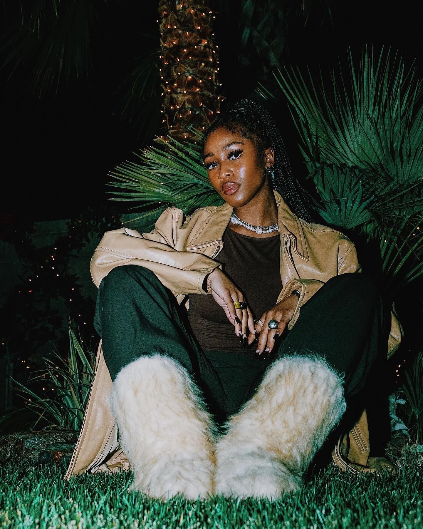 A woman sitting outdoors at night among lush greenery, wearing a beige jacket, black pants, and white furry boots, with jewelry including rings, a necklace, and earrings, illuminated by string lights in the background.