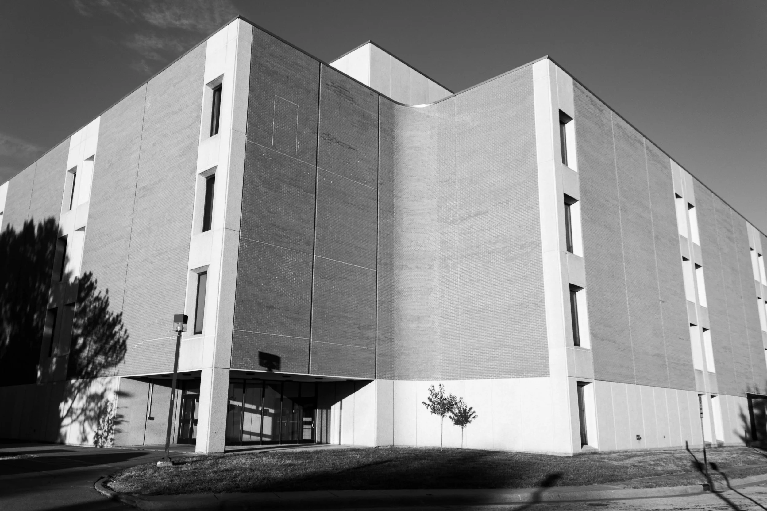 Black and white photo of a modern multi-story building with a curved corner, small trees in front, and a sidewalk in the foreground.