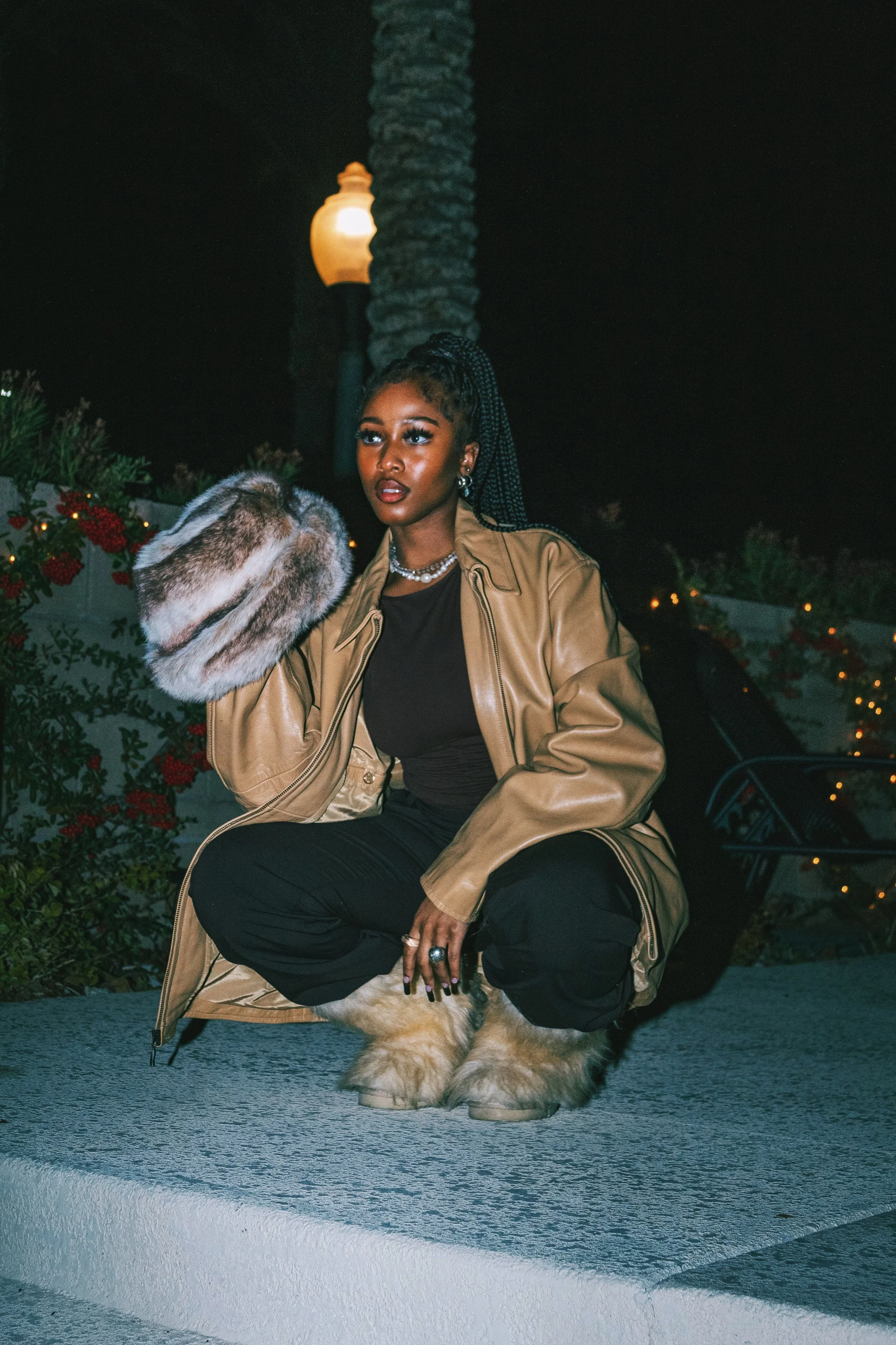 A woman crouching outdoors at night, wearing a tan leather oversized jacket, black shirt and pants, fur boots, holding a fur hat, with bushes and decorative lights behind her.