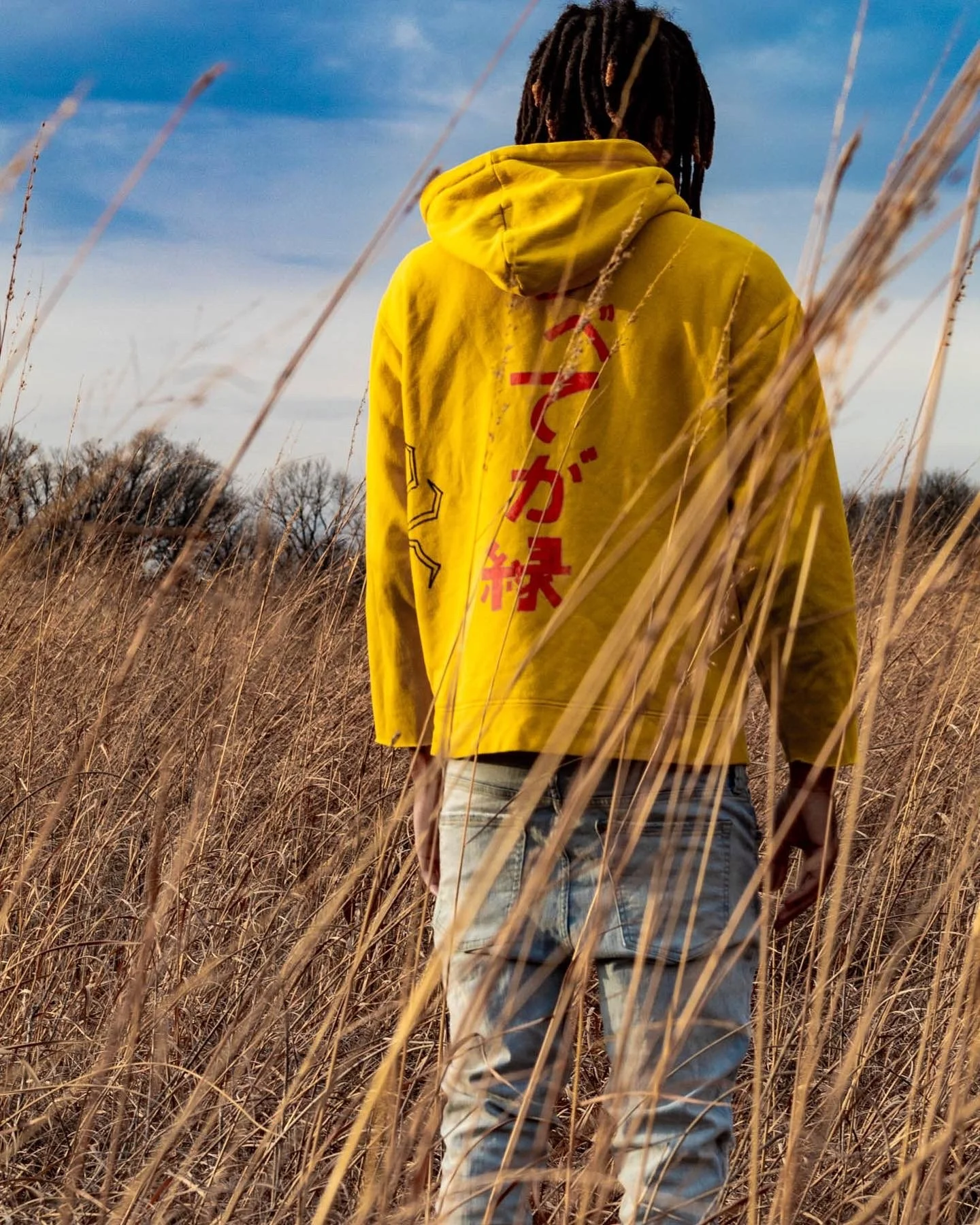 Person with dreadlocks wearing a yellow hoodie with red Japanese characters on the back, standing in a field of tall dry grass with a cloudy sky in the background.