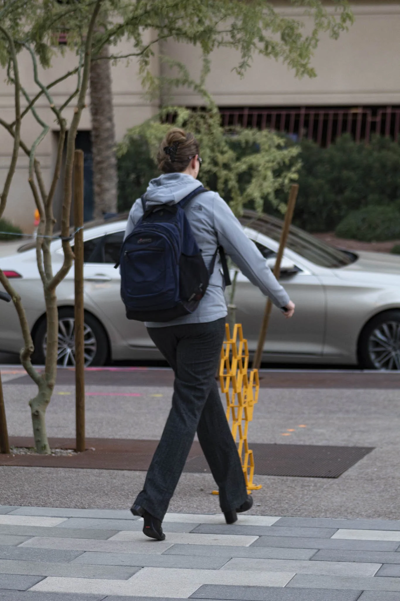 A woman with a backpack walking across a crosswalk on a city street, carrying two yellow folding stools.