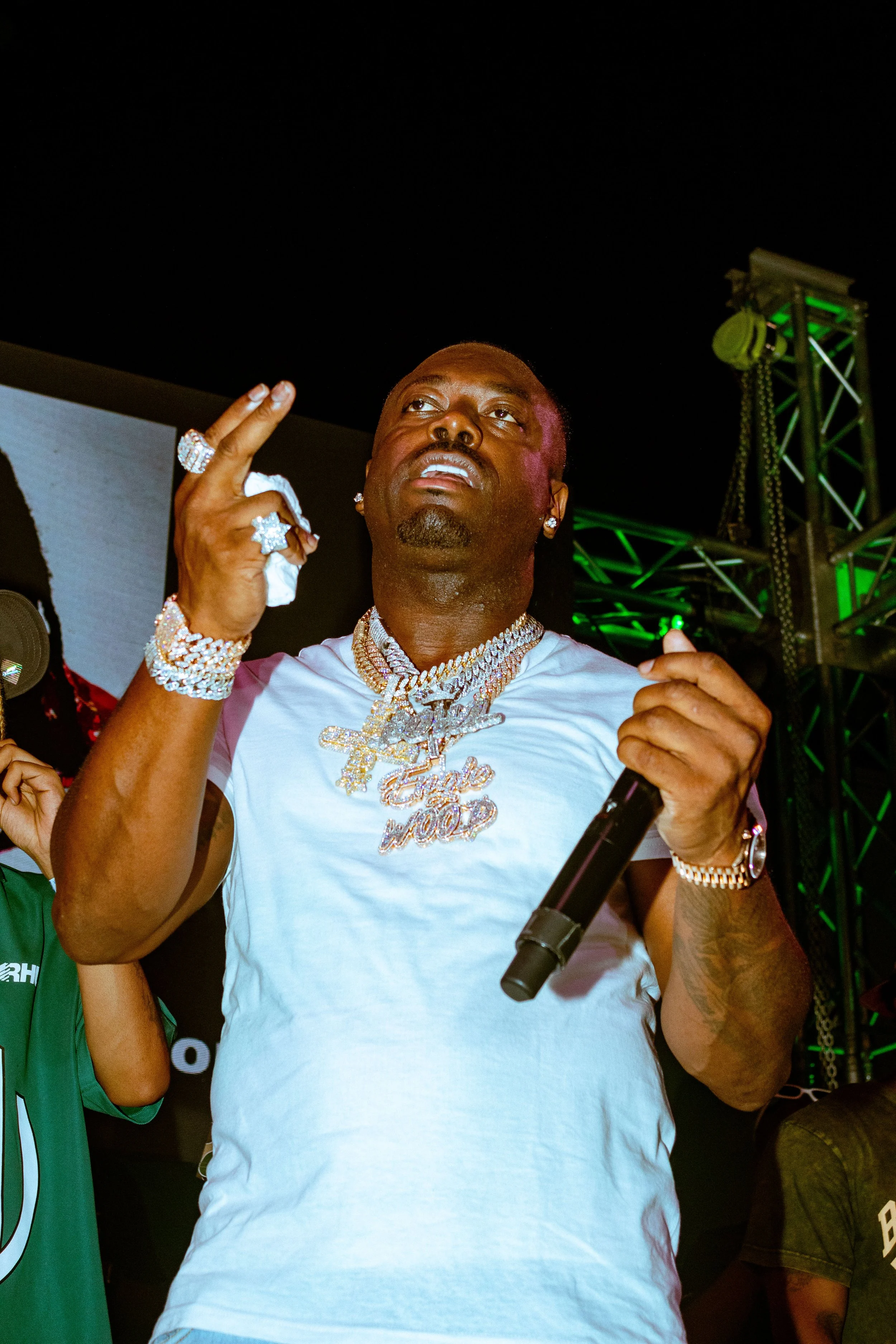 A man wearing a white T-shirt and multiple diamond jewelry, holding a microphone and making a gesture with his hand, at a nighttime event with stage lighting.
