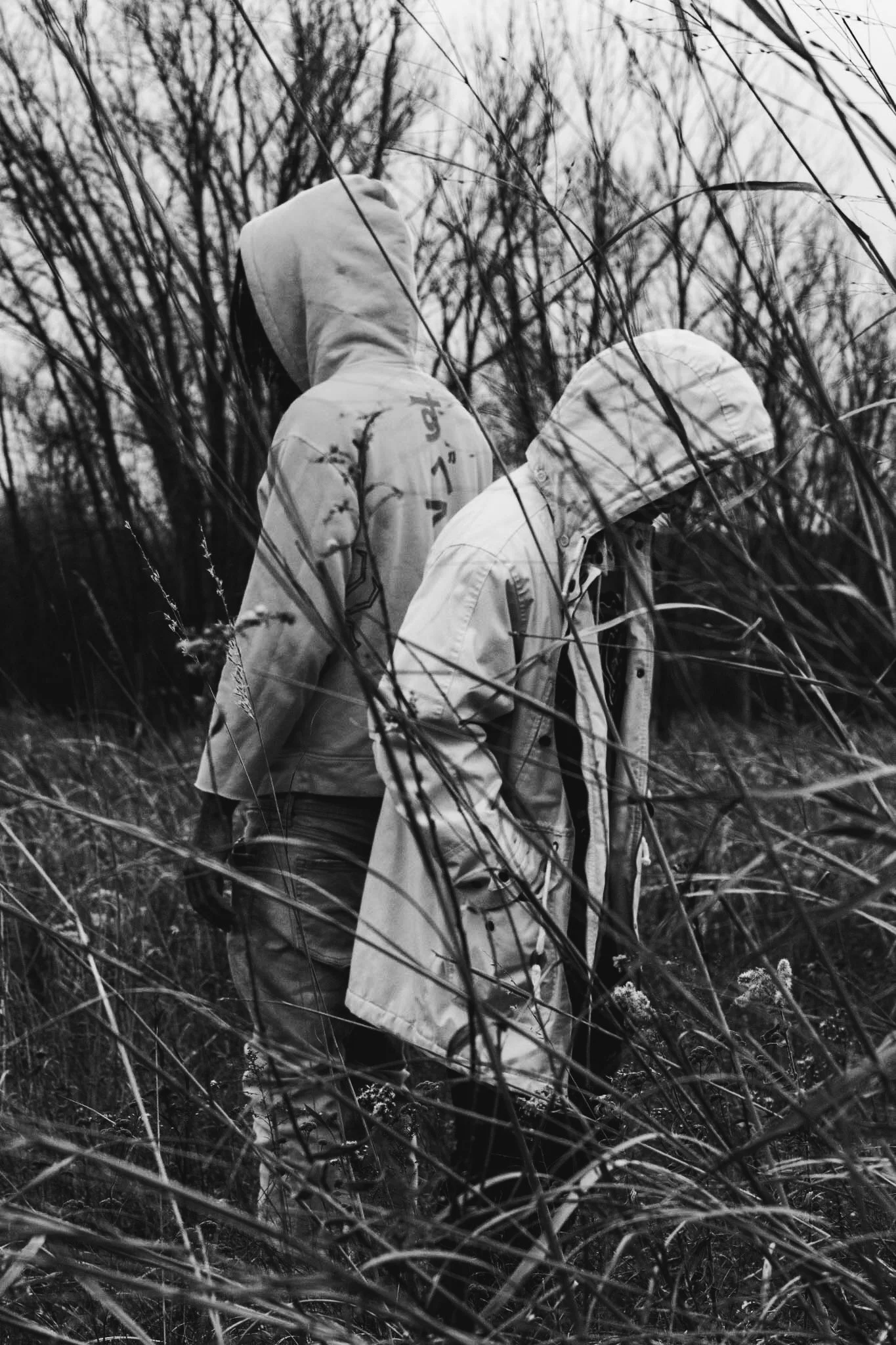 Two people wearing hooded jackets walking through tall grass and leafless trees outdoors during overcast weather.