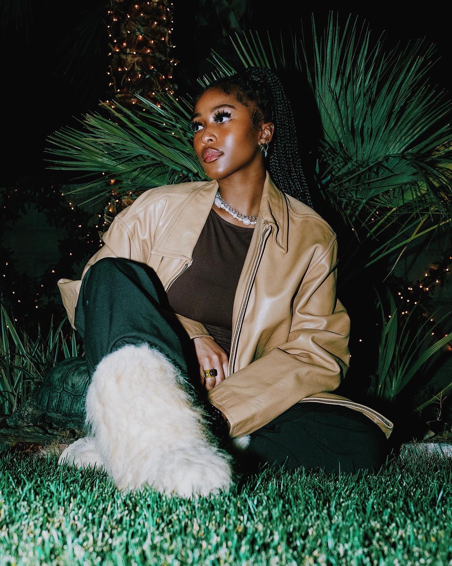 A young Black woman with long braided hair, sitting outdoors at night, surrounded by greenery and string lights, wearing a tan leather jacket, black pants, and fluffy white boots, looking thoughtfully into the distance.
