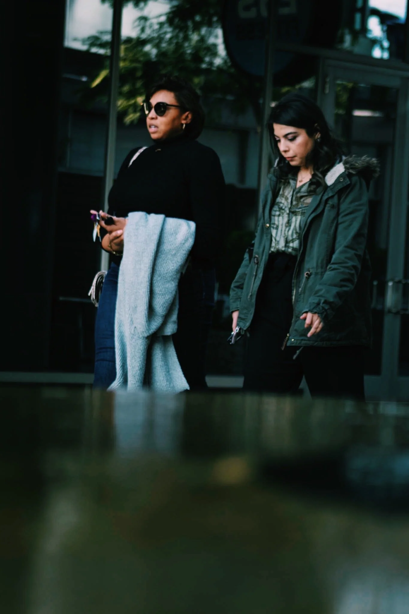 Two women walking outside, one wearing sunglasses and holding keys, the other looking down, both wearing jackets.