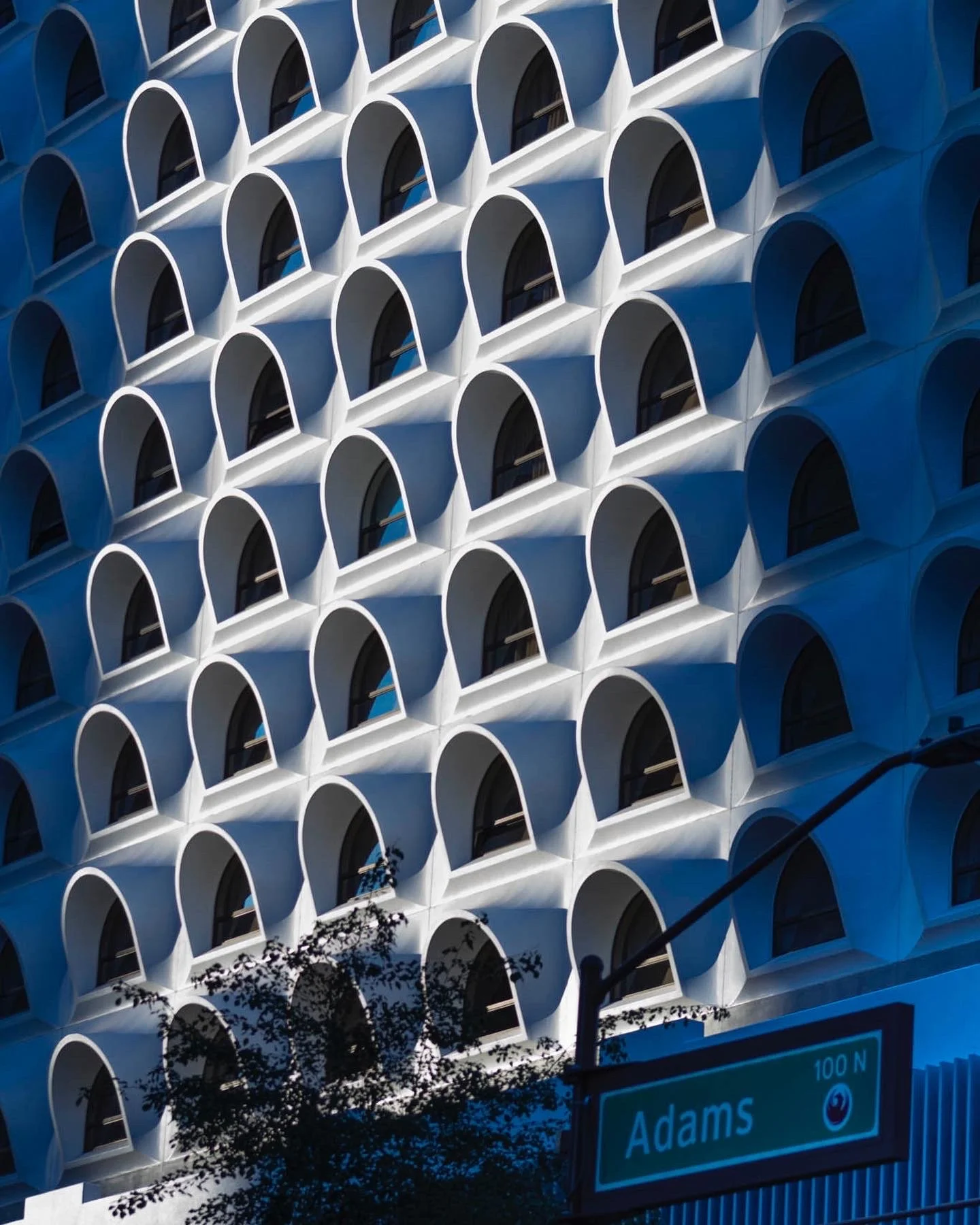 Close-up of a modern building's facade with repetitive white arched balconies and shadows, and part of a street sign reading 'Adams' with a street light.