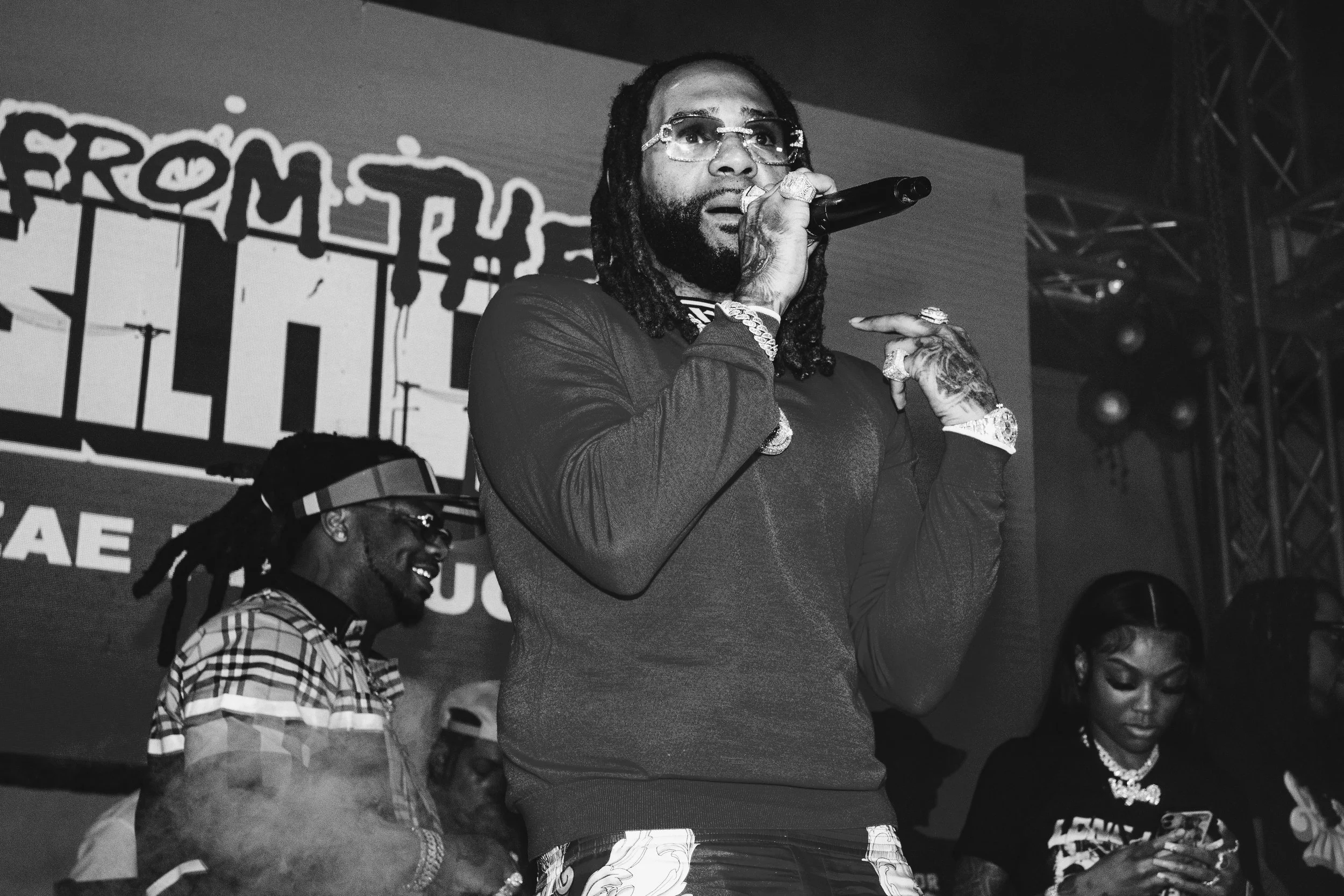 A man with dreadlocks, sunglasses, and jewelry holding a microphone on stage, with a large screen and other people behind him.