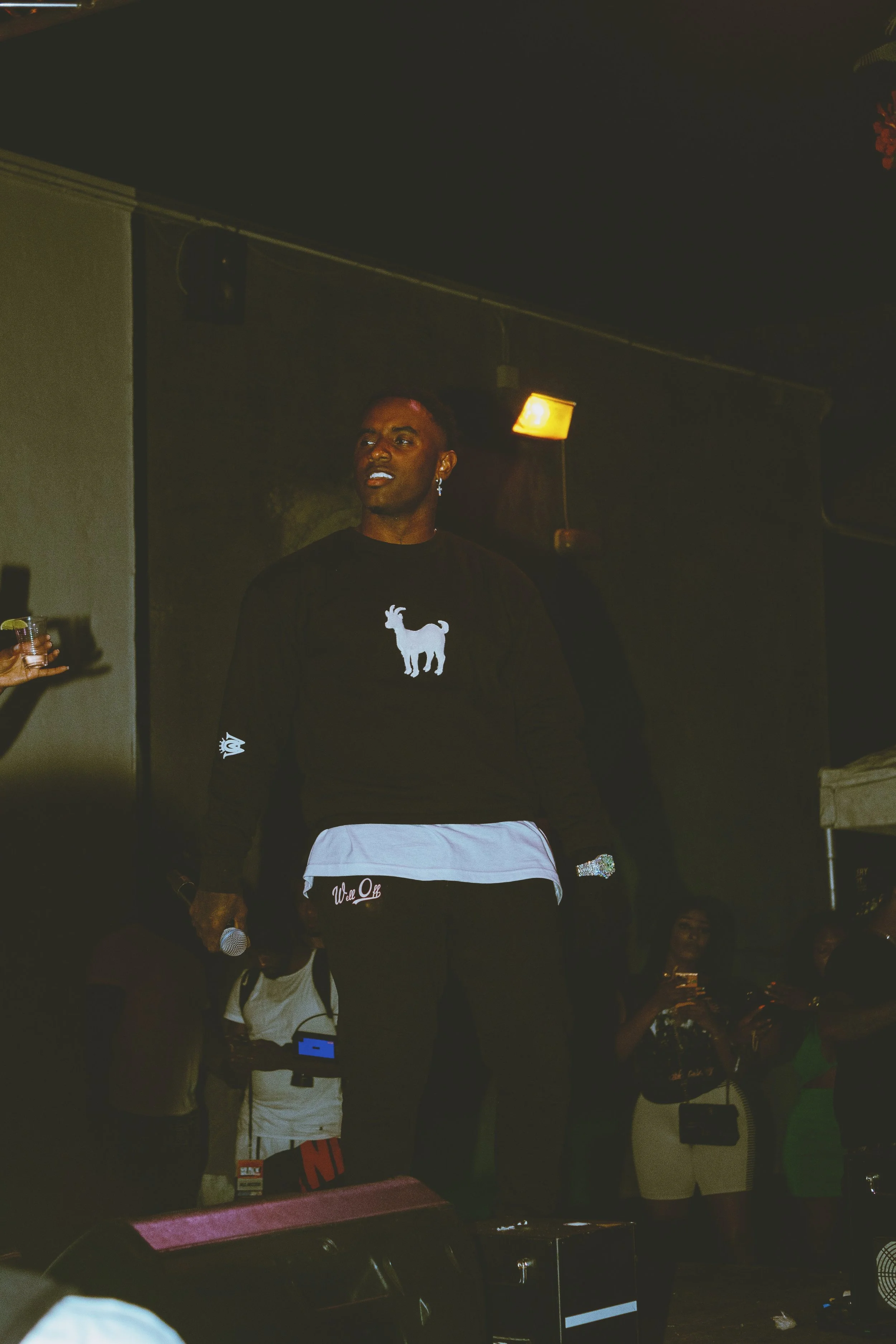 A man standing on a stage holding a microphone, wearing a black sweater with a white goat logo, and several people in the background at a social event.