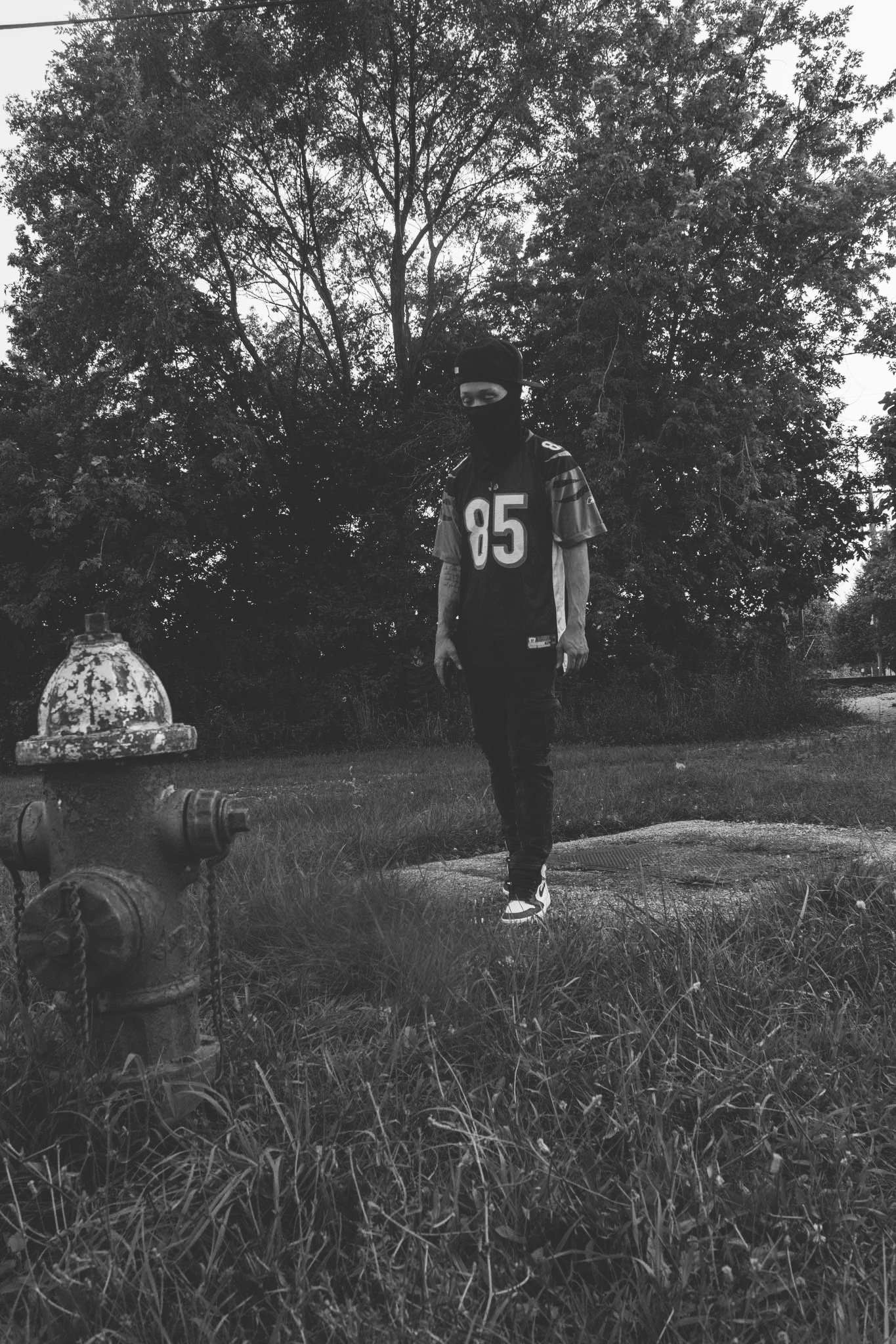 A person in a black mask and sports jersey with the number 85 stands near a fire hydrant in an outdoor park, with trees in the background.