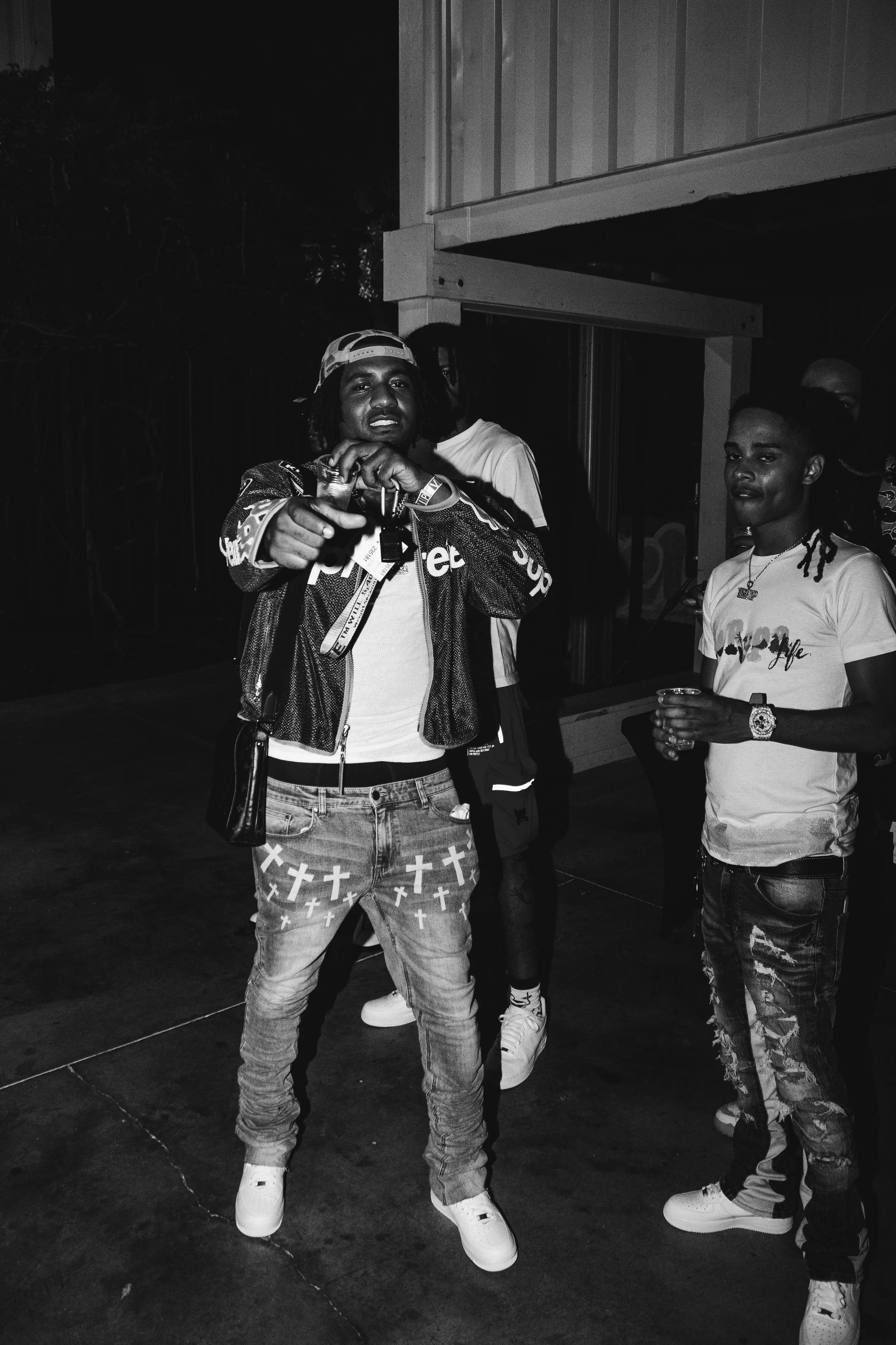 Group of young men socializing outdoors at night, dressed in casual streetwear, with one person holding a drink and wearing a cap backwards.