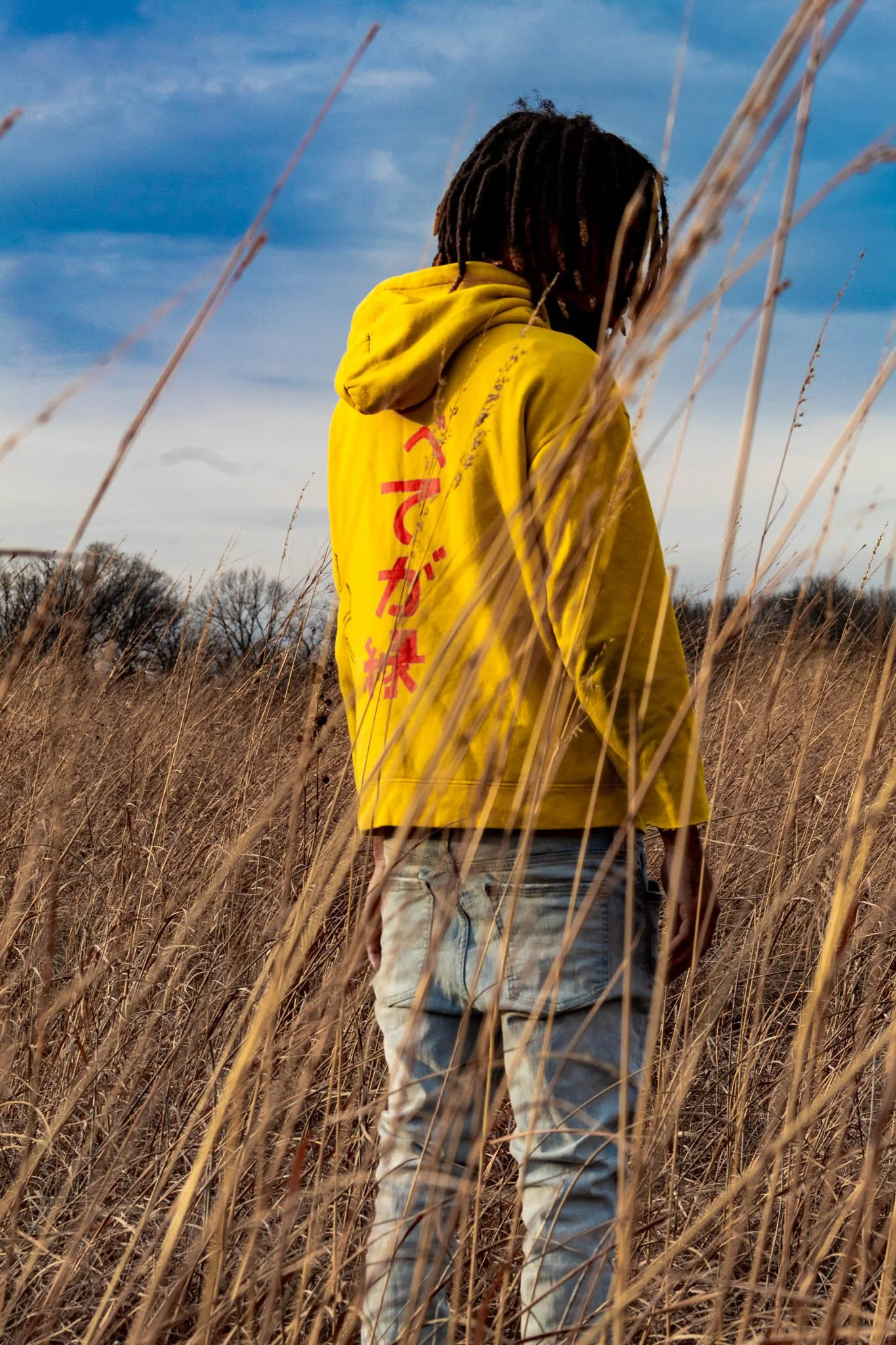 Person with dreadlocks wearing a yellow hoodie and distressed jeans standing in a field of tall dry grass under a partly cloudy sky.