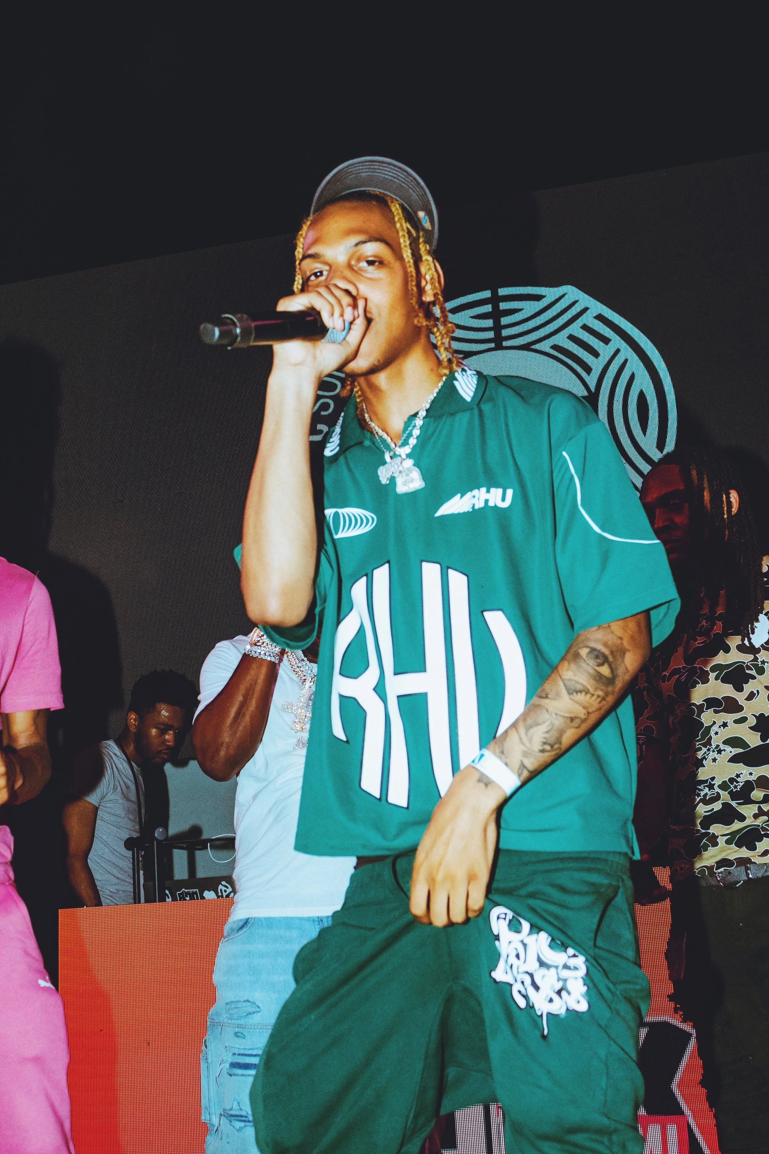 A young man with blond dreadlocks wearing a green sports jersey and matching pants, holding a microphone on stage at a music event.