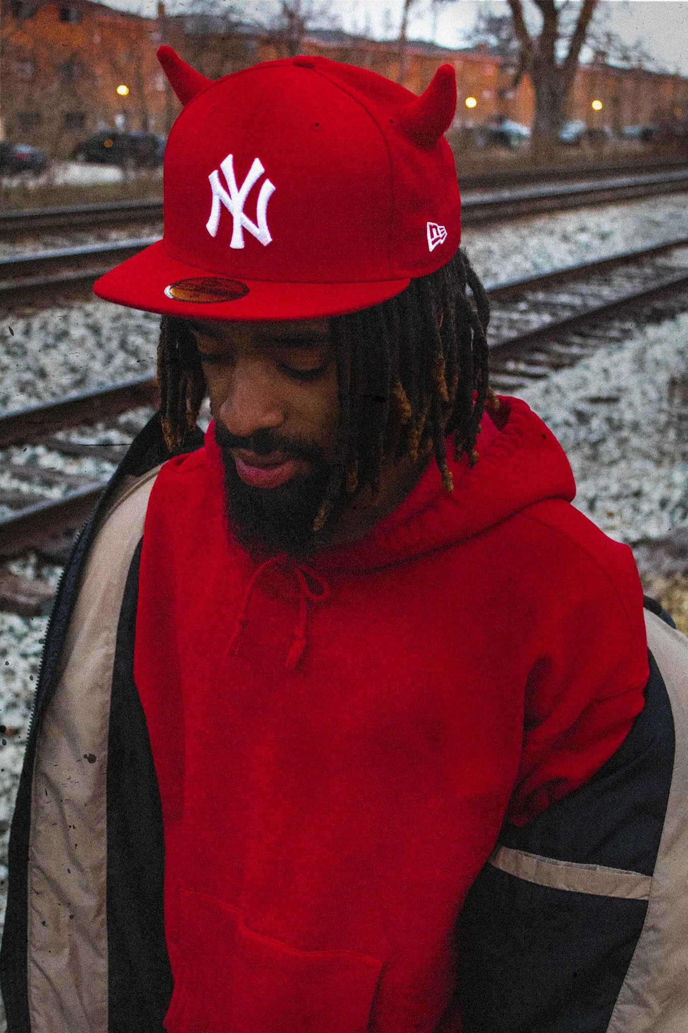 Man with dreadlocks wearing a red hoodie and a red New York Yankees cap with devil horns, standing near train tracks outdoors at dusk.
