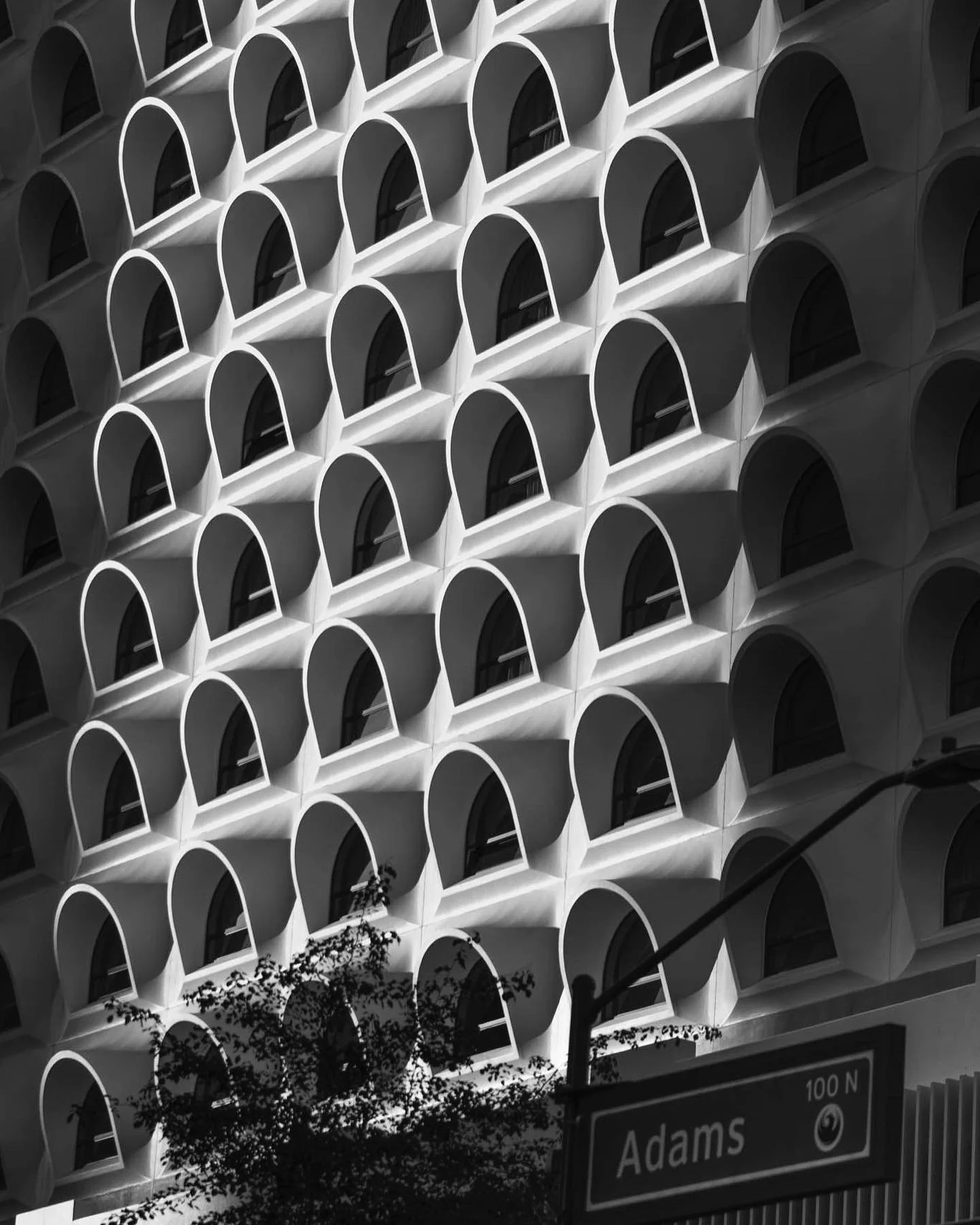 Black and white photo of a modern building with numerous identical arched windows and a street sign that reads 'Adams 100 N'.