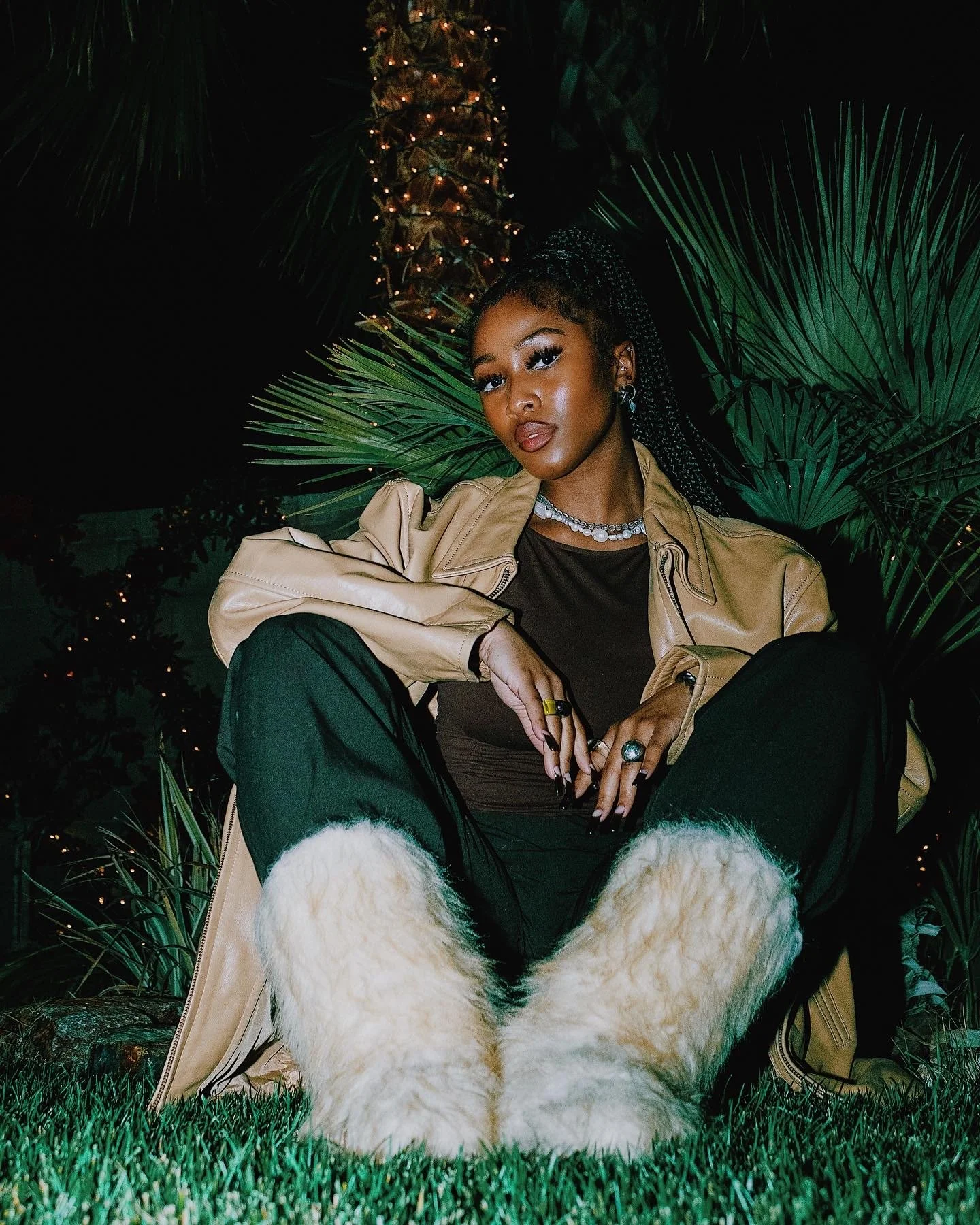 A woman sitting outdoors at night surrounded by palm leaves, wearing a beige jacket, black pants, and fluffy white boots, with jewelry and makeup, illuminated in a dark setting with decorative lights.