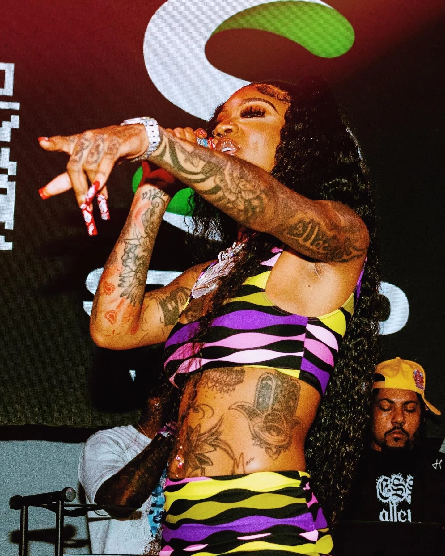 A woman with tattoos and long curly black hair performing on stage, wearing a colorful striped crop top and matching skirt, with a DJ and another person visible in the background.