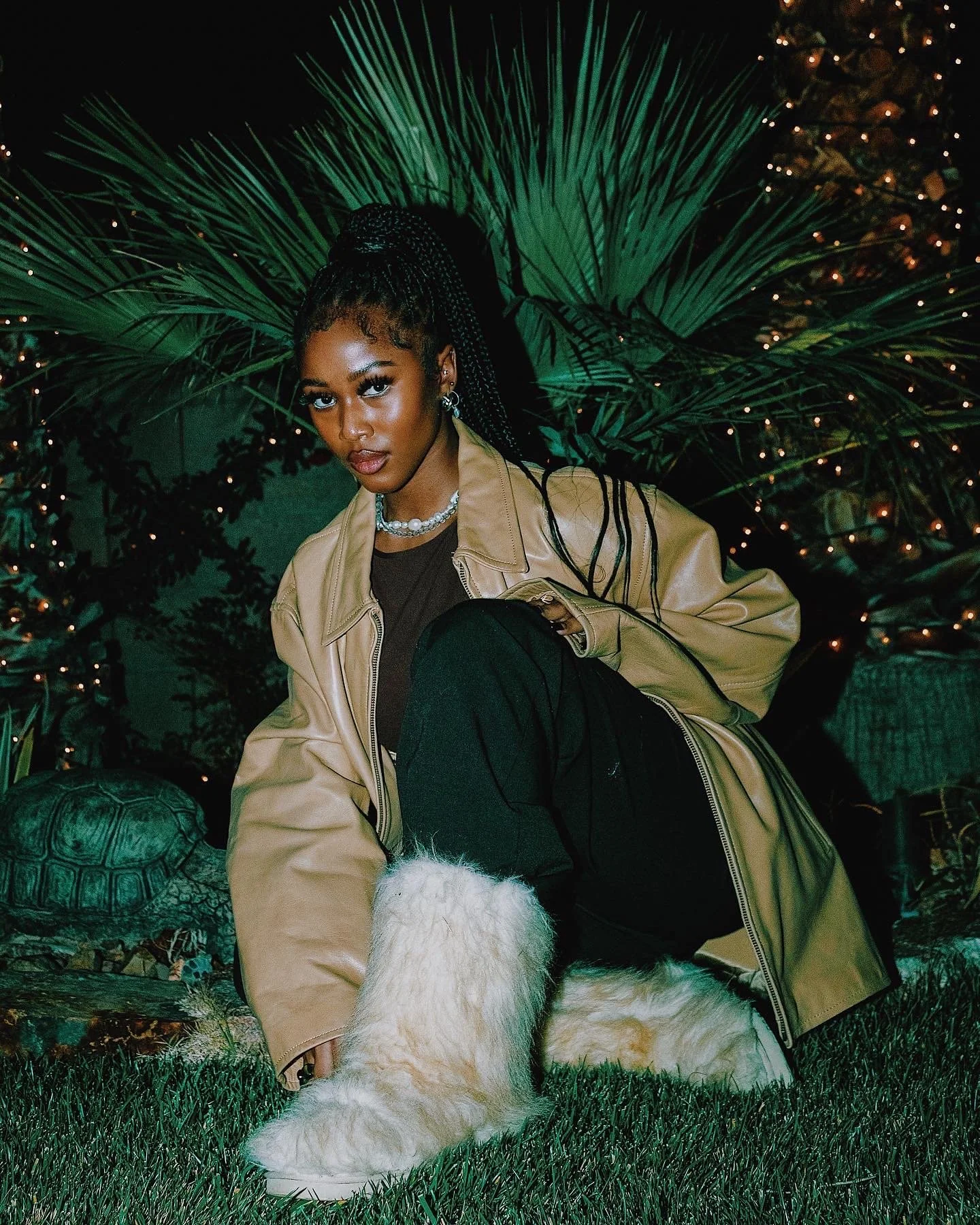 A young woman with braided hair poses outdoors at night surrounded by lush green plants and twinkling string lights. She wears a beige jacket, black clothing, white furry boots, and jewelry.