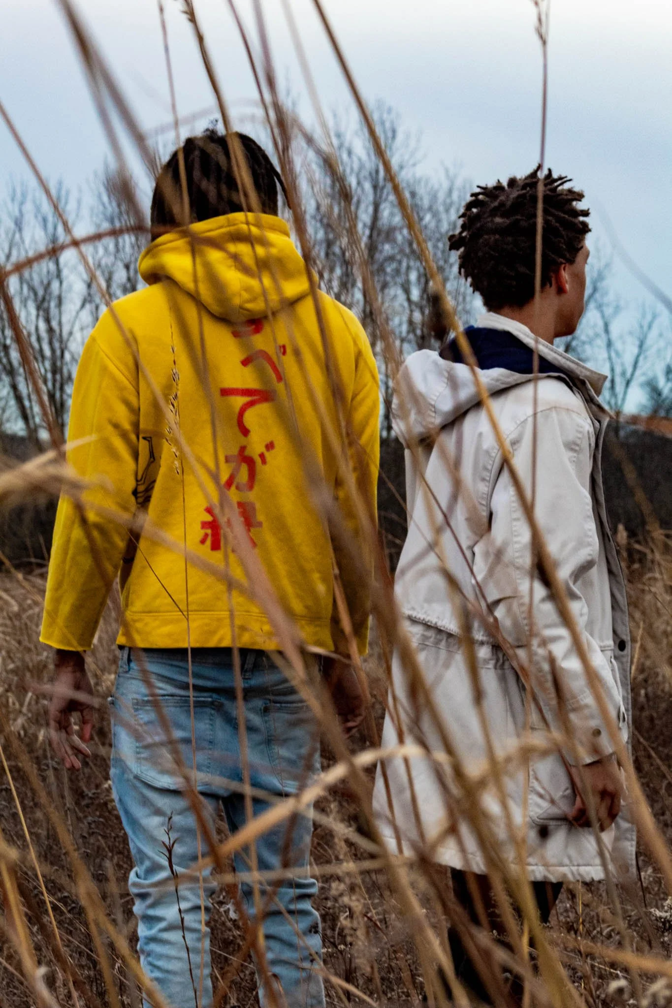 Two young men walking through a field of tall, dried grass on a cloudy day. One is wearing a bright yellow hoodie with Japanese characters on the back, and the other is wearing a light-colored jacket. Bare trees are visible in the background.