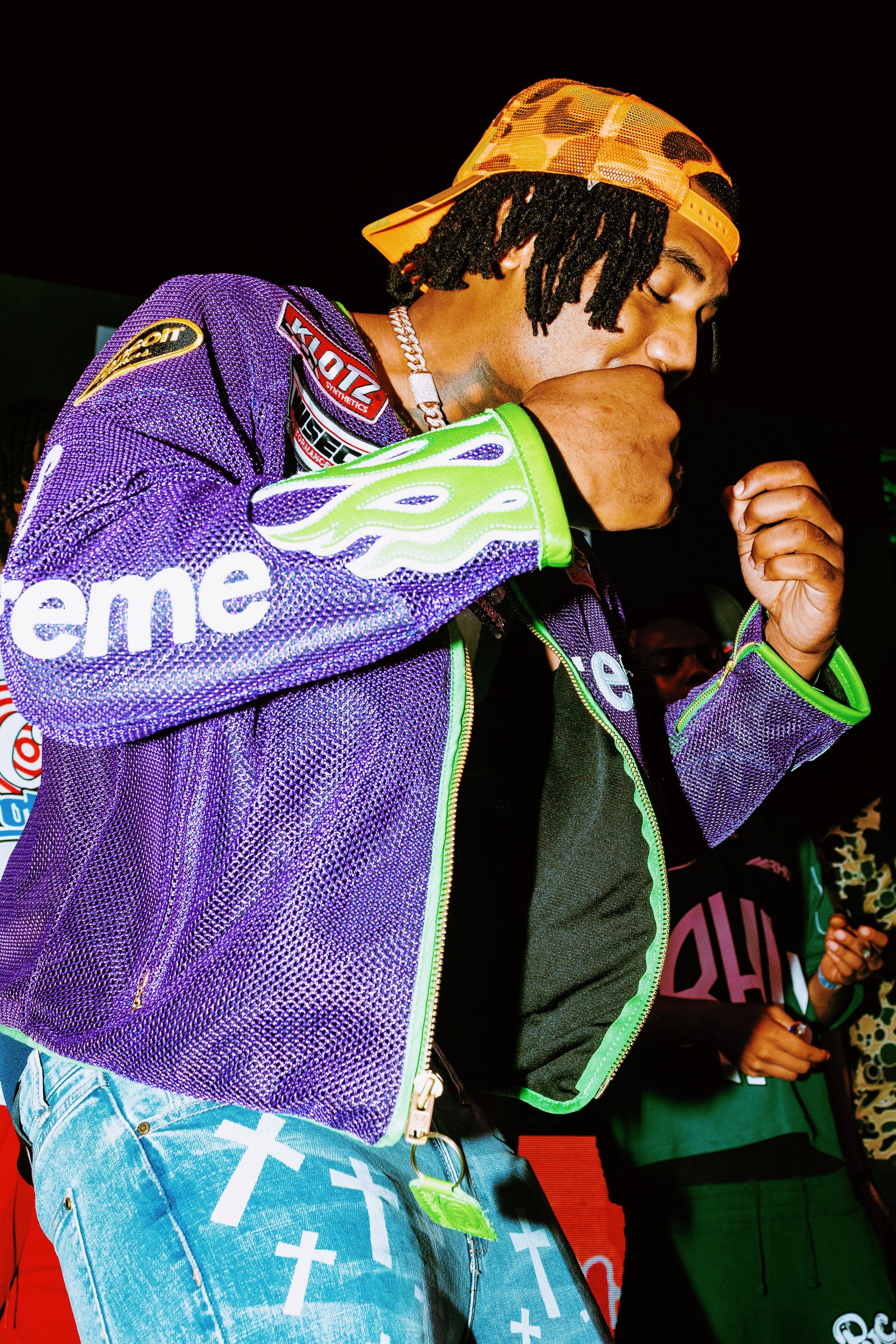 A young man wearing a purple mesh jacket with logos and patches, a black shirt, light blue jeans with cross patterns, and a yellow and orange camo cap backward, is in a close-up shot with his eyes closed, holding his hand near his mouth.