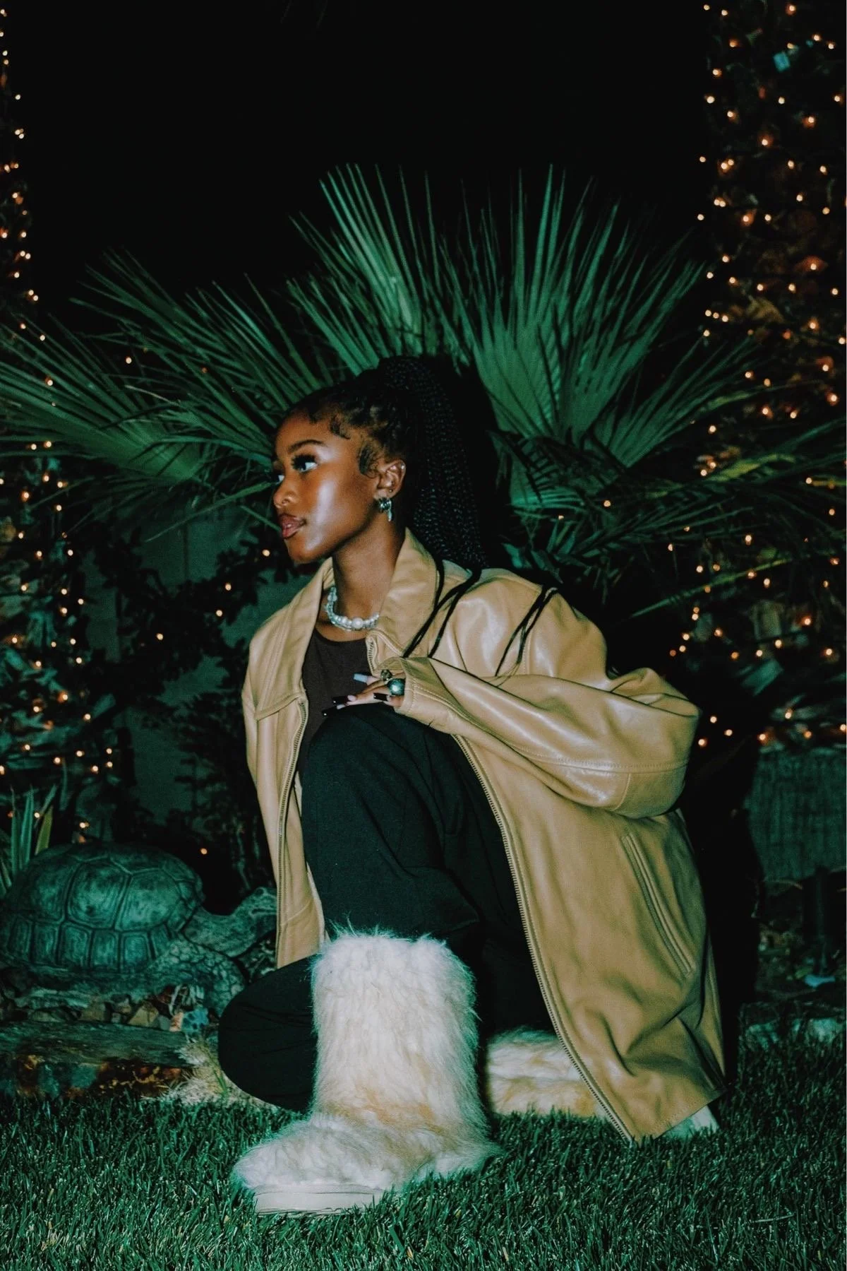A woman with braided hair wearing a beige leather jacket, black pants, and white furry boots, kneeling on grass at night with a backdrop of palm leaves and Christmas trees decorated with lights.