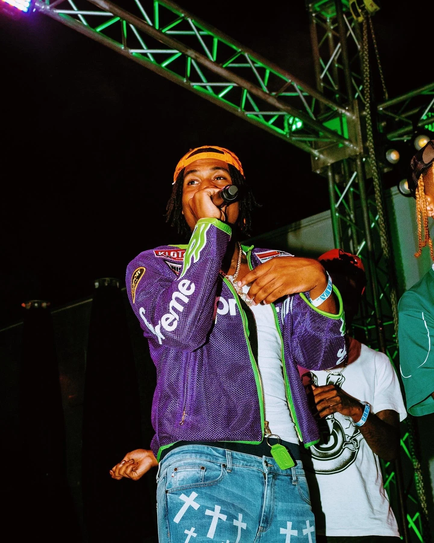 Person wearing purple Supreme jacket and jeans with top crosses, speaking into a microphone on stage with stage lighting and metal truss in background.