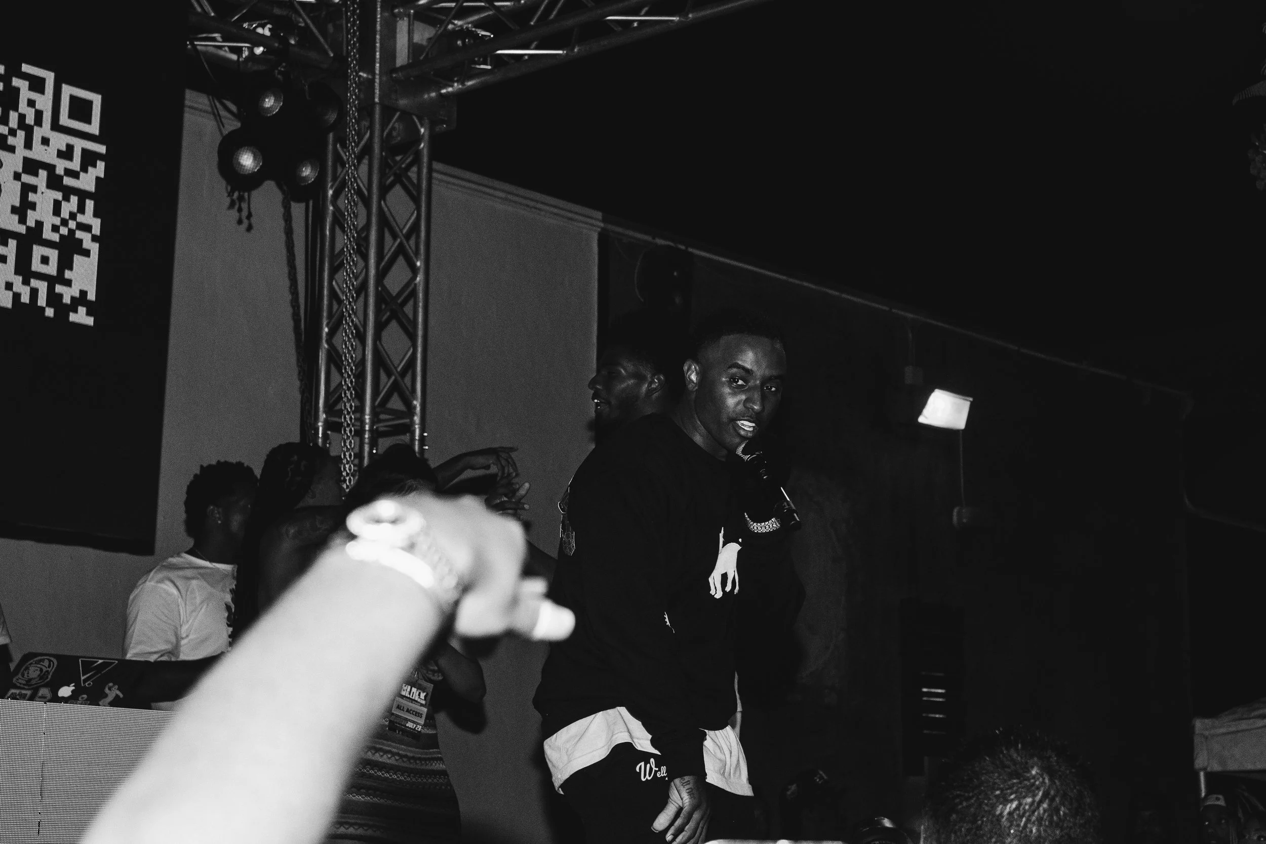 A man performing on stage with a microphone in hand, engaging the crowd, with others dancing behind him in a dark venue.