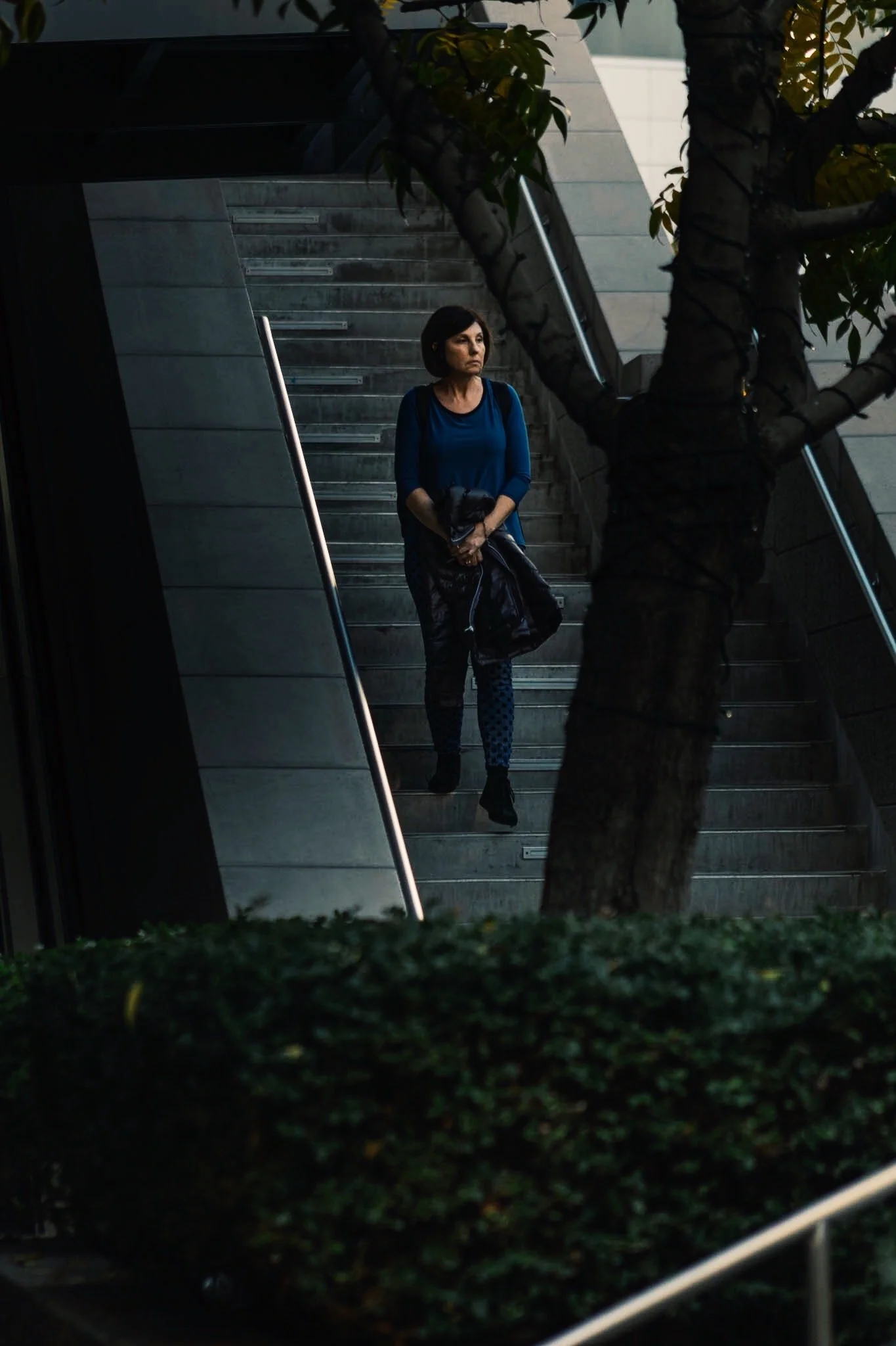 A woman with short dark hair, wearing a blue top and patterned pants, is walking down a set of stairs outside, holding a black jacket and a bag, partially obscured by a tree in the foreground.