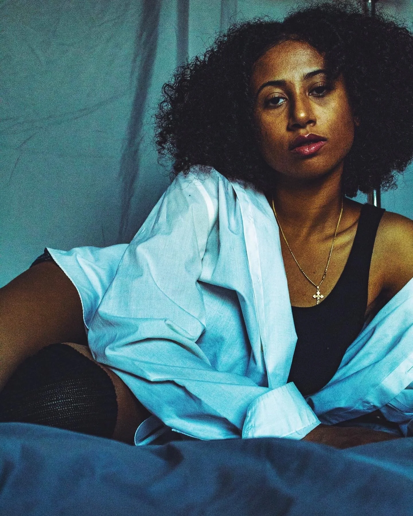 A woman with curly hair and medium skin tone wearing a black tank top, a gold necklace with a cross pendant, and a light blue shirt. She is sitting on a bed with a dark background, looking at the camera with a serious expression.