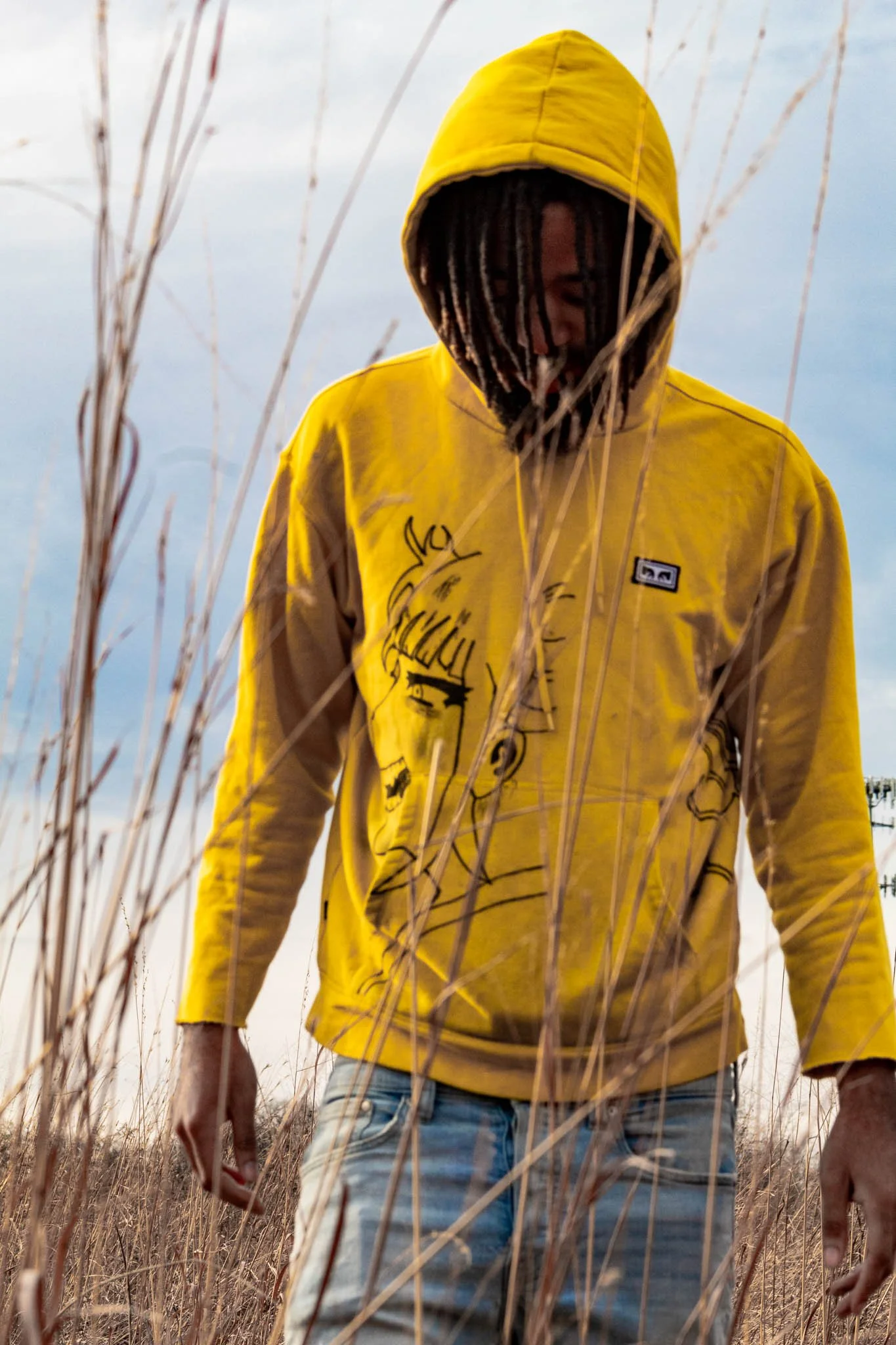 A person wearing a yellow hoodie with artwork of a girl's face, standing in tall, dry grass with an overcast sky in the background.