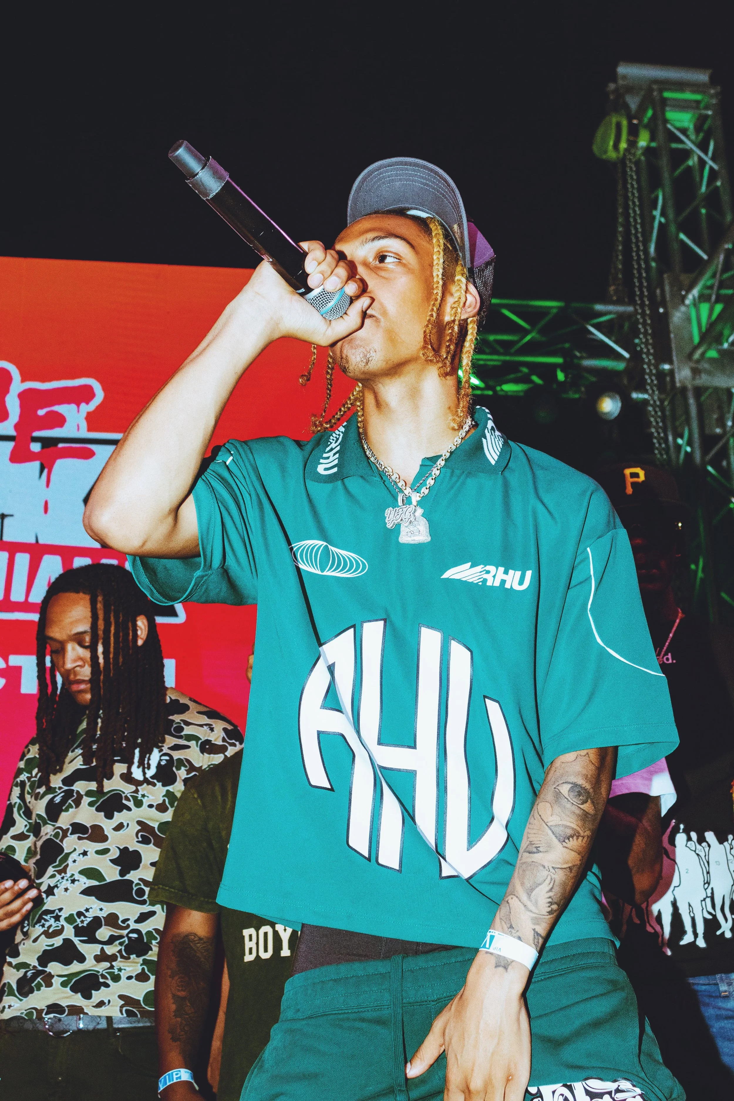 A male performer with braids and jewelry singing into a microphone at a concert stage, with intense lighting and a red background.