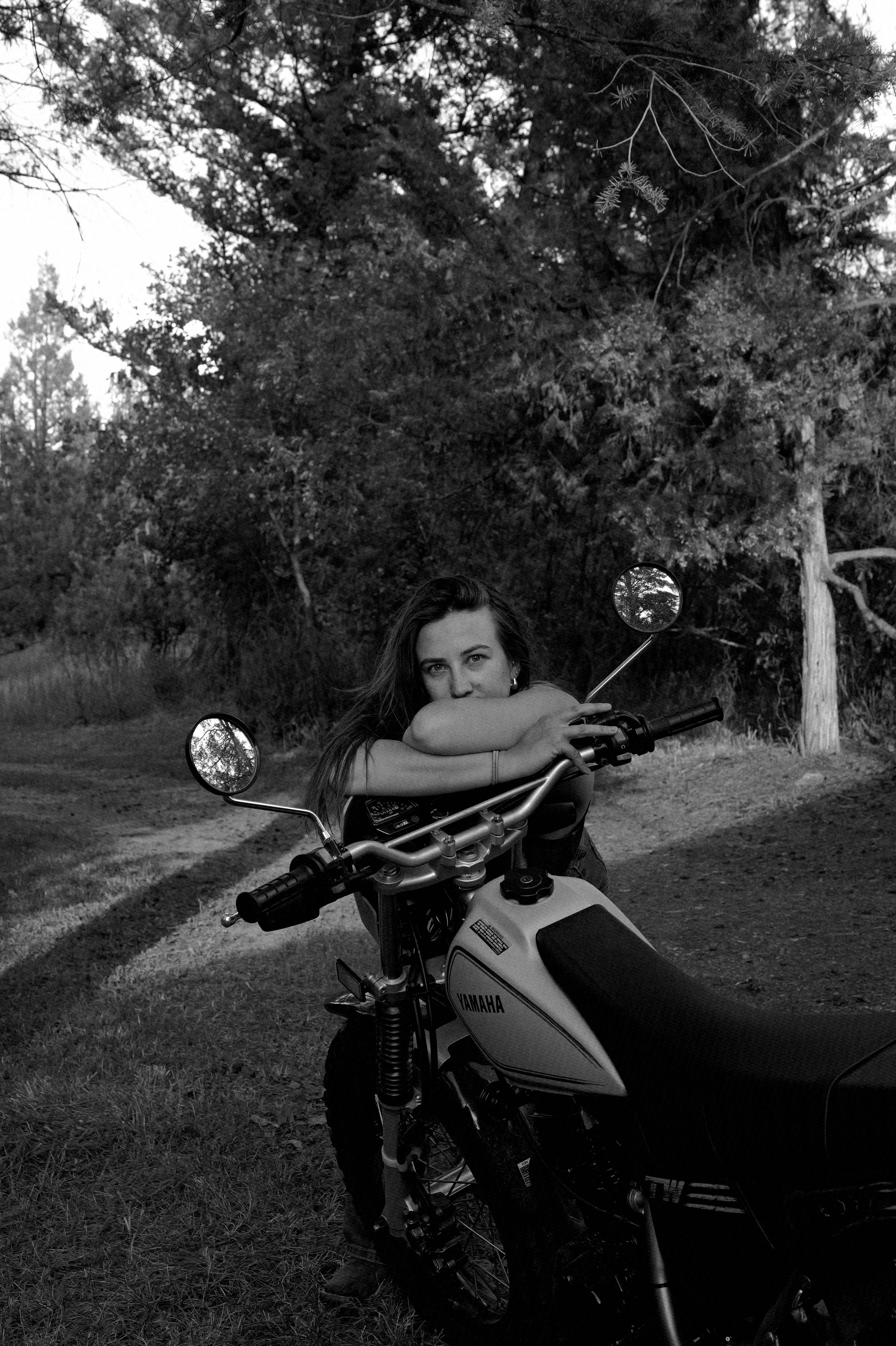 A woman leaning on a Yamaha motorcycle on a dirt path surrounded by trees.