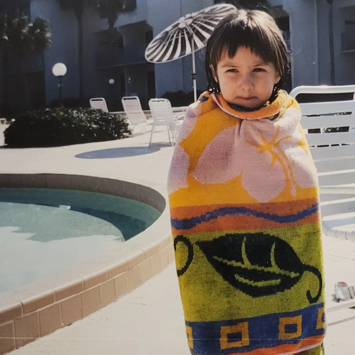 Young child wrapped in a colorful beach towel standing next to a swimming pool on a sunny day.