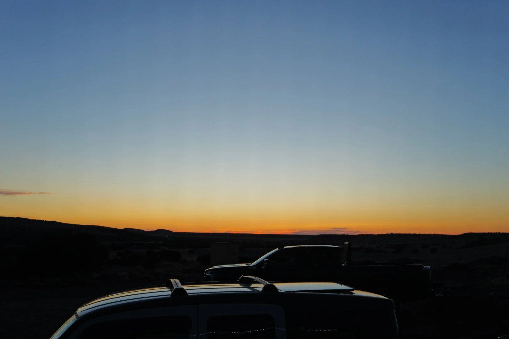 A sunset or sunrise with two trucks parked in a landscape with hills in the distance.