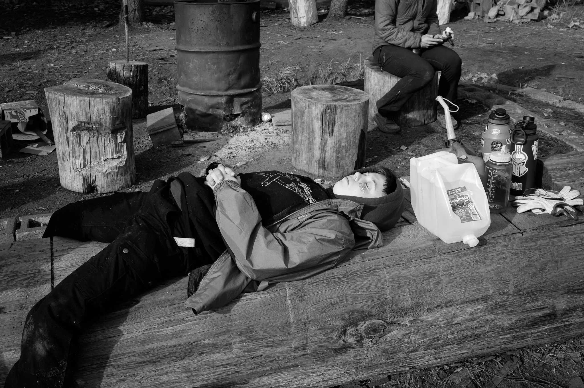 A person lying on a large wooden log outdoors, with eyes closed and hands clasped over their stomach, surrounded by various bottles, containers, gloves, and other items. In the background, another person is sitting on a tree stump, using a mobile device.
