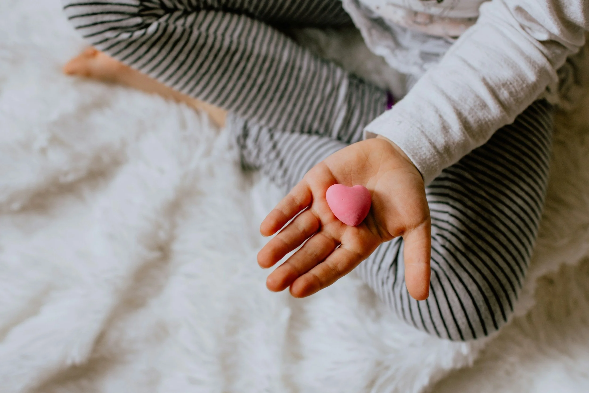 Child sitting on a white fuzzy blanket, holding a pink heart-shaped object in their hand.