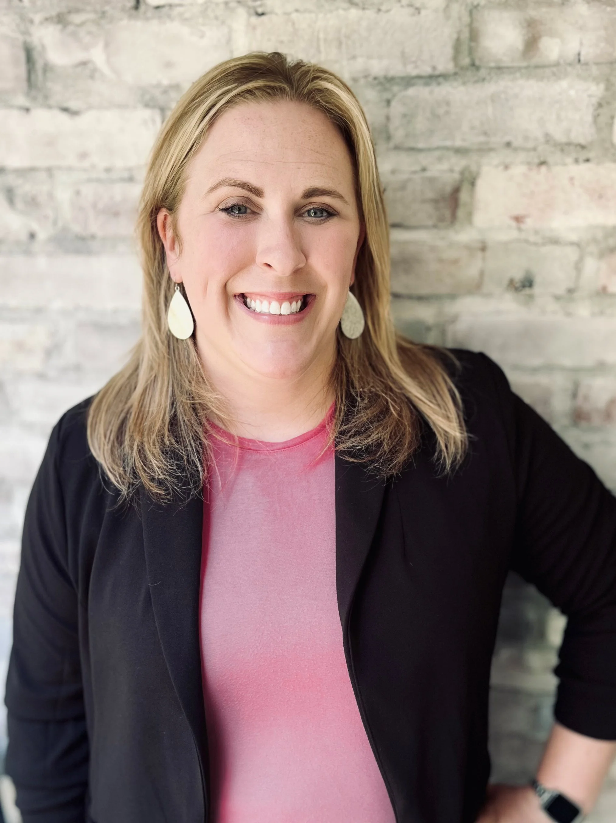 A photograph of Emily who has blonde hair and is wearing a black blazer, pink top, and large teardrop earrings, while standing against a stone wall.